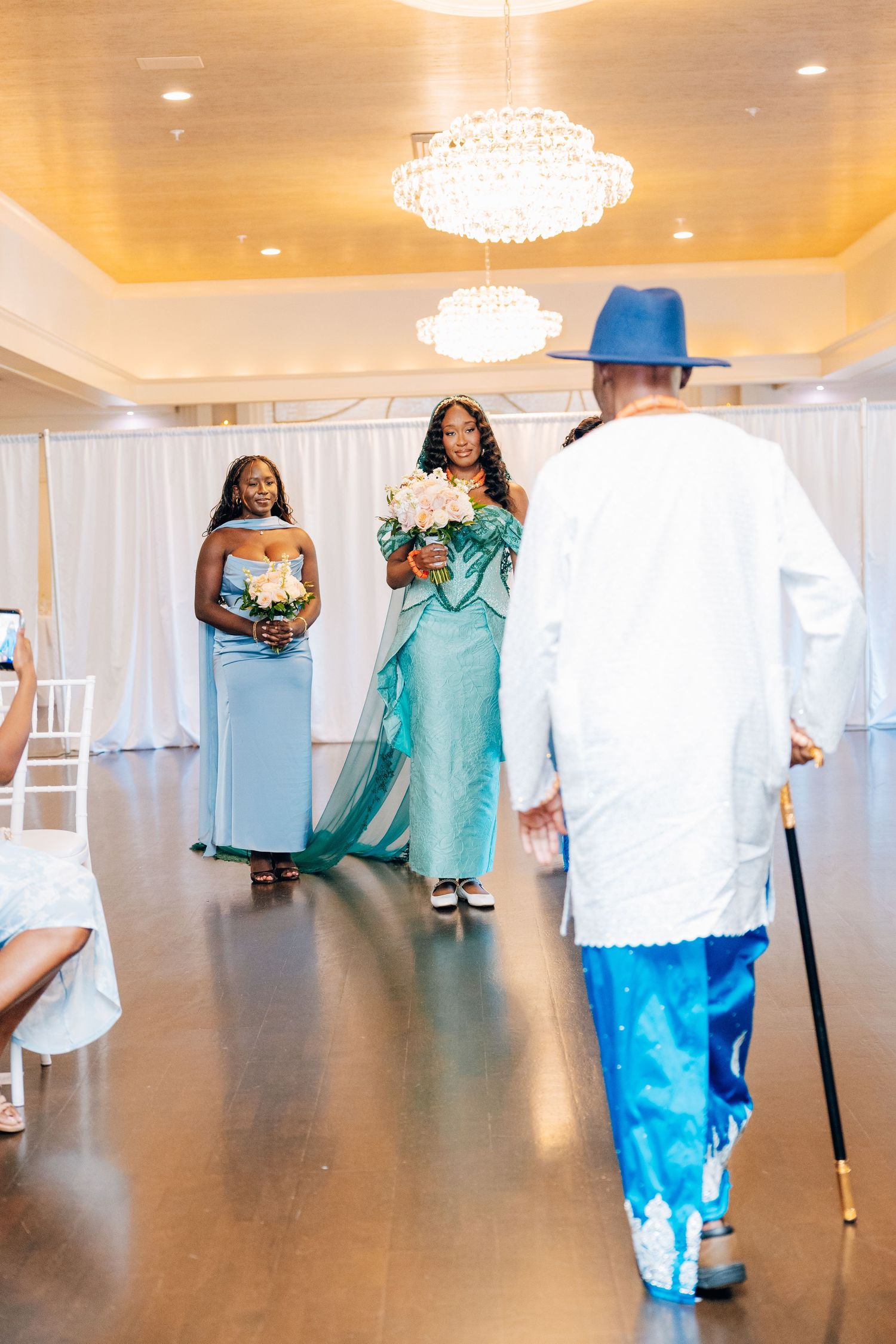 Bride in teal African gown walks down aisle toward elder man in traditional blue outfit.