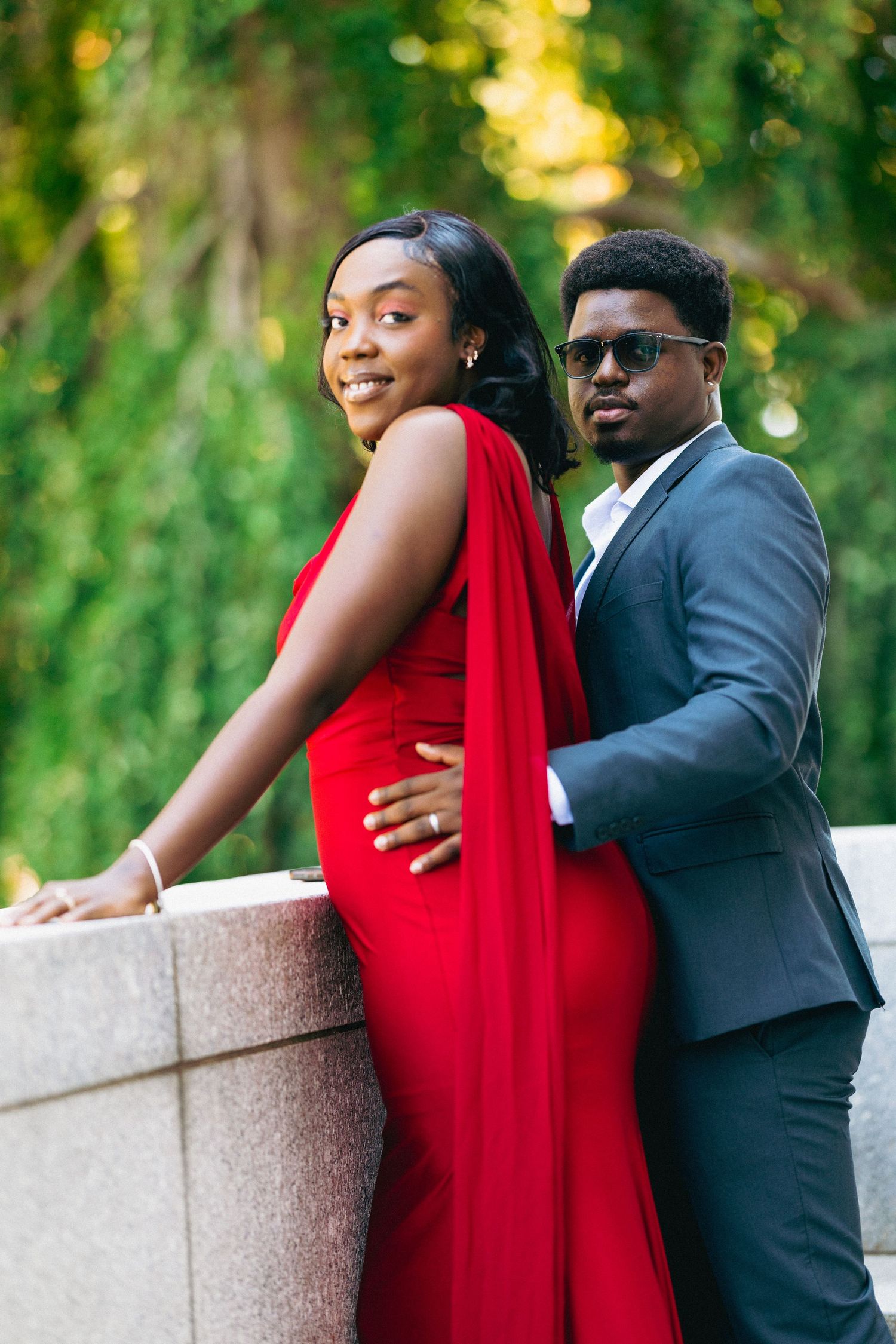 A smiling woman in a red gown and a man in a gray suit pose back-to-back against a stone wall outdoors.