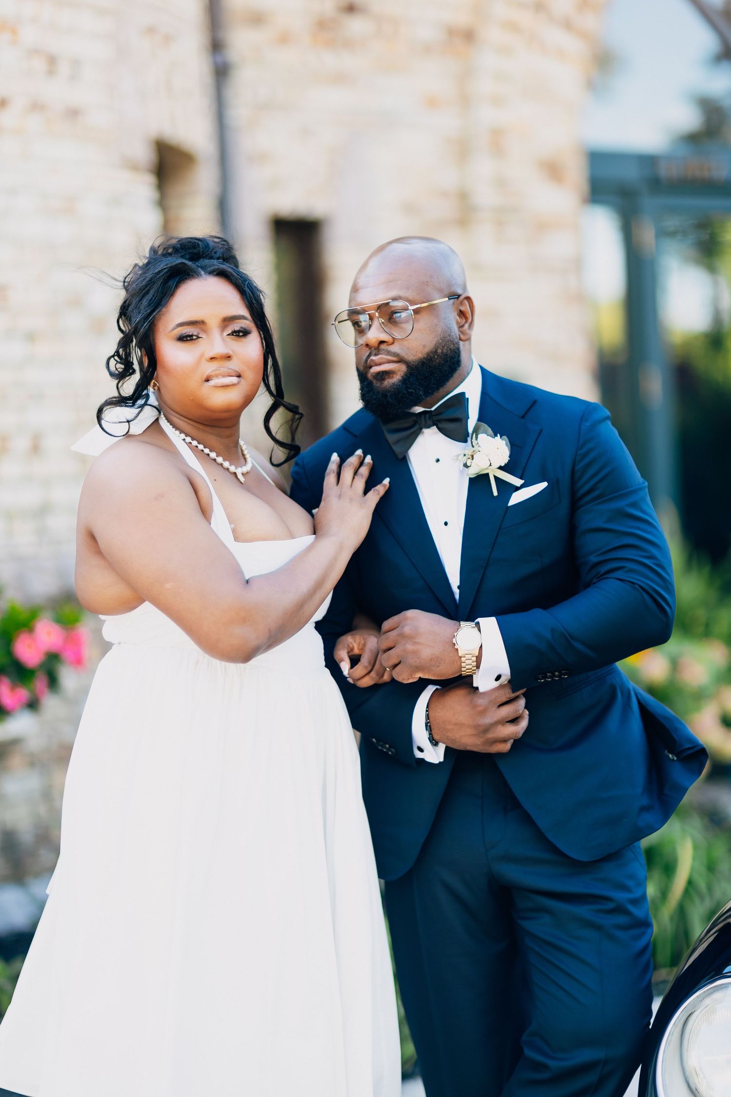 Bride and groom posing elegantly outdoors, groom in navy tuxedo with boutonniere, bride in white halter gown with pearl necklace.