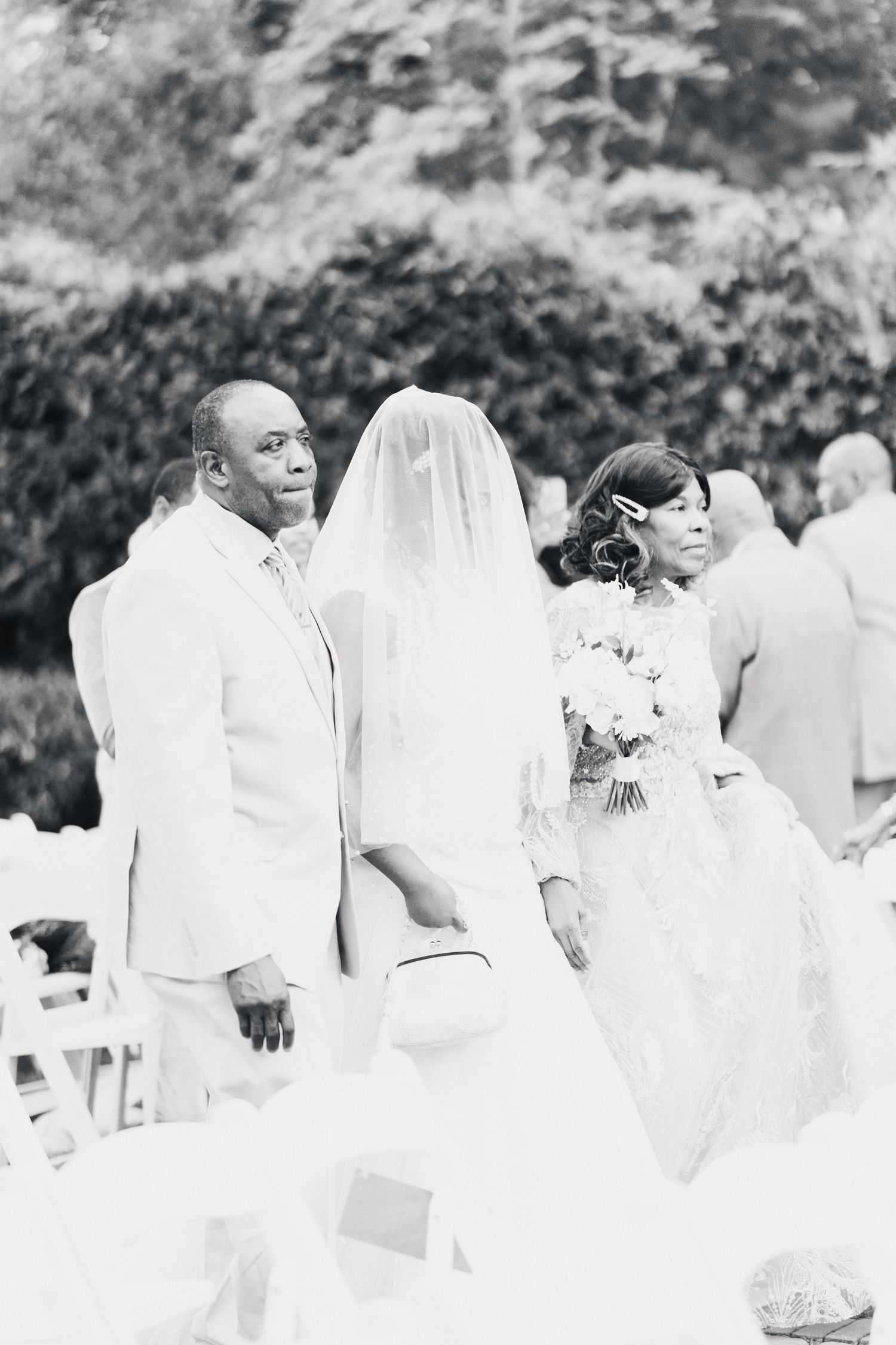 Black and white outdoor wedding ceremony with bride in veil and gown, groom in white suit, and guests seated.