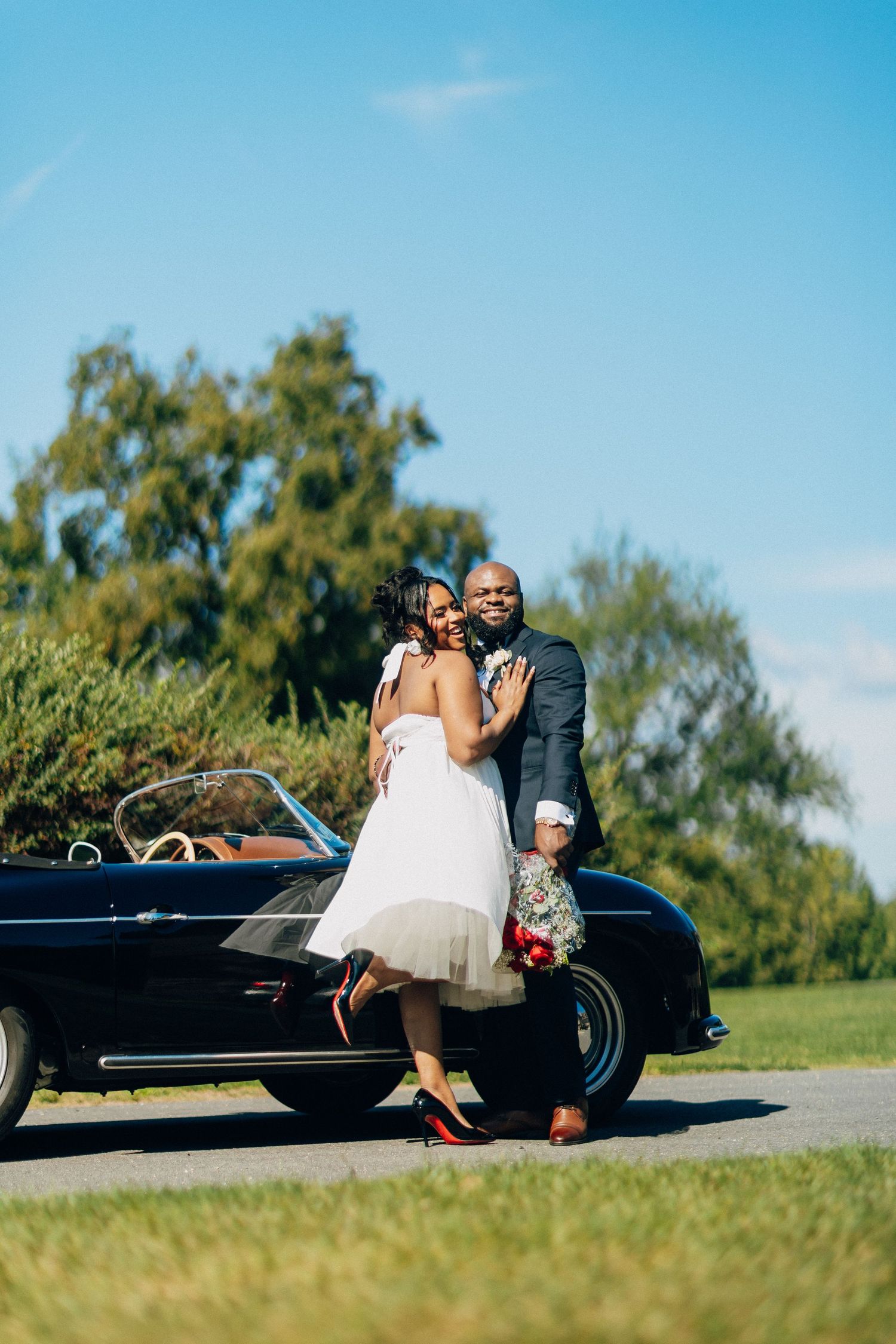Black bride and groom embracing beside a classic navy blue convertible on a sunny outdoor path.