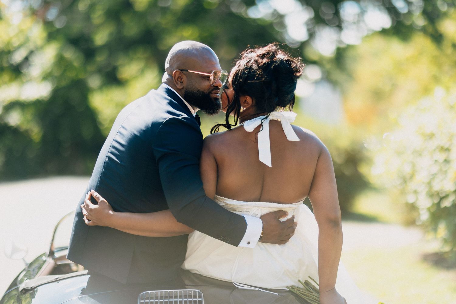 Groom kisses bride on the cheek while seated together in a vintage convertible outdoors.