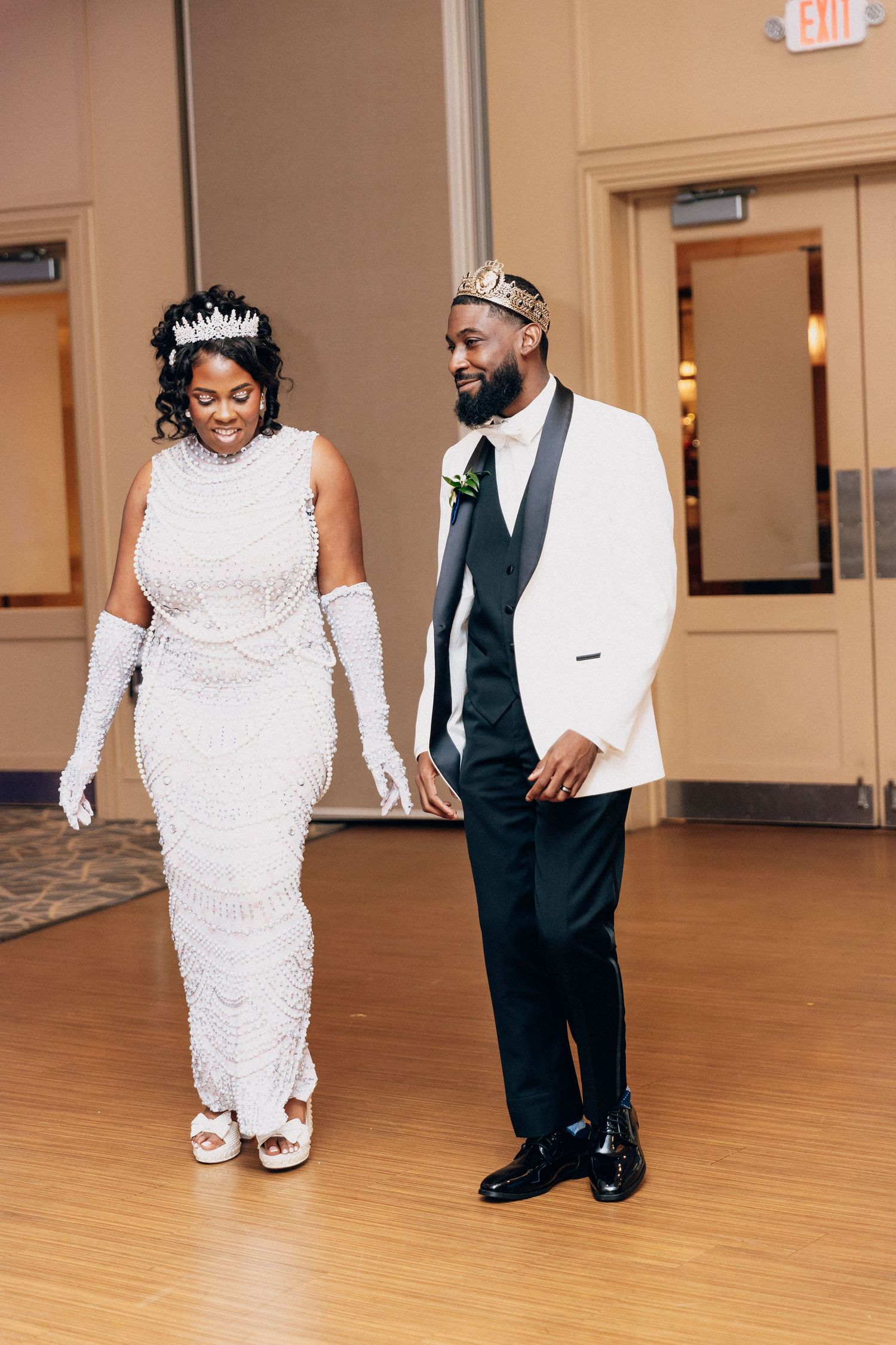 Newlyweds wearing crowns walk hand in hand at their wedding reception in matching white outfits.