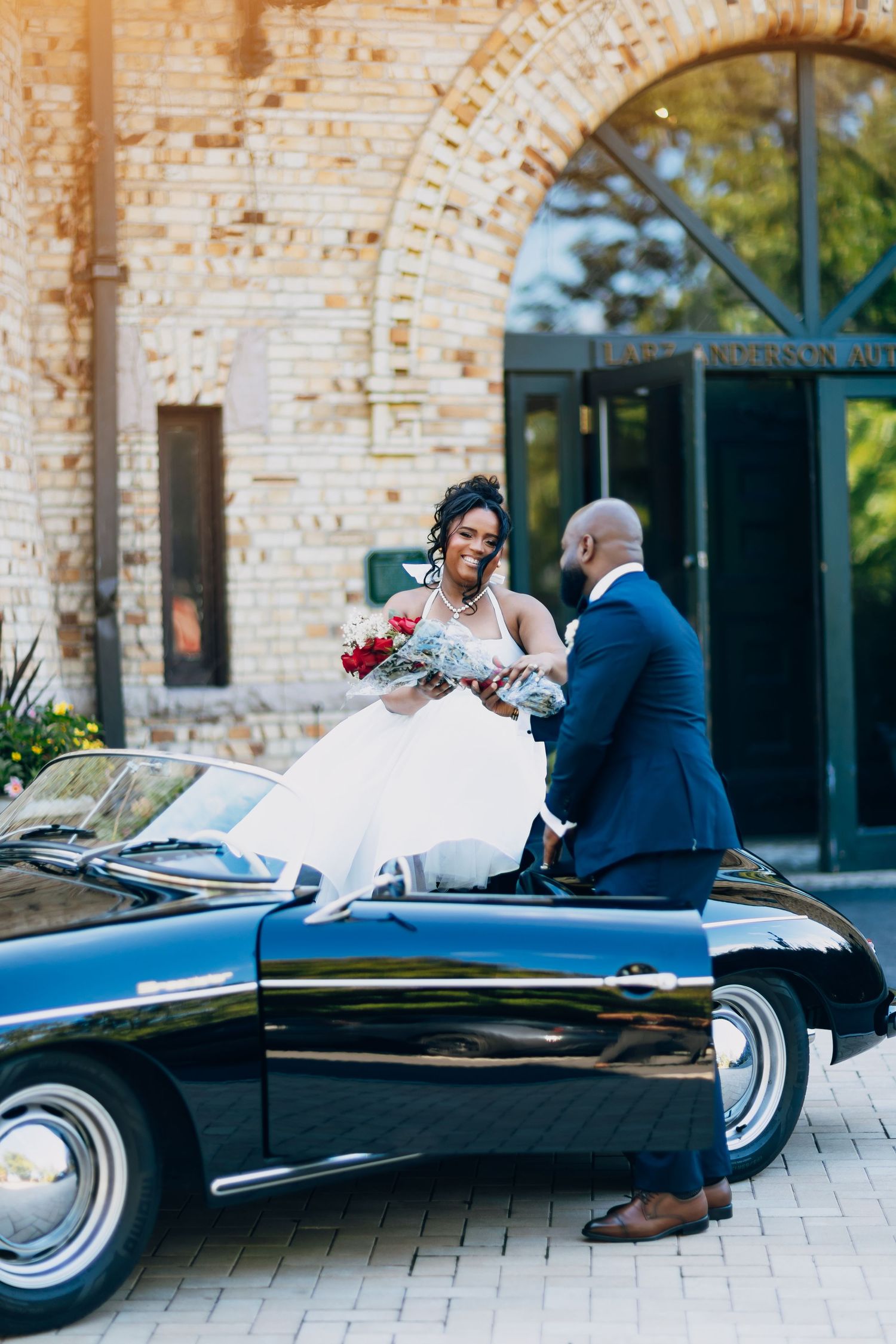 Smiling bride holding red bouquet stepping out of a vintage black Porsche as groom assists her.
