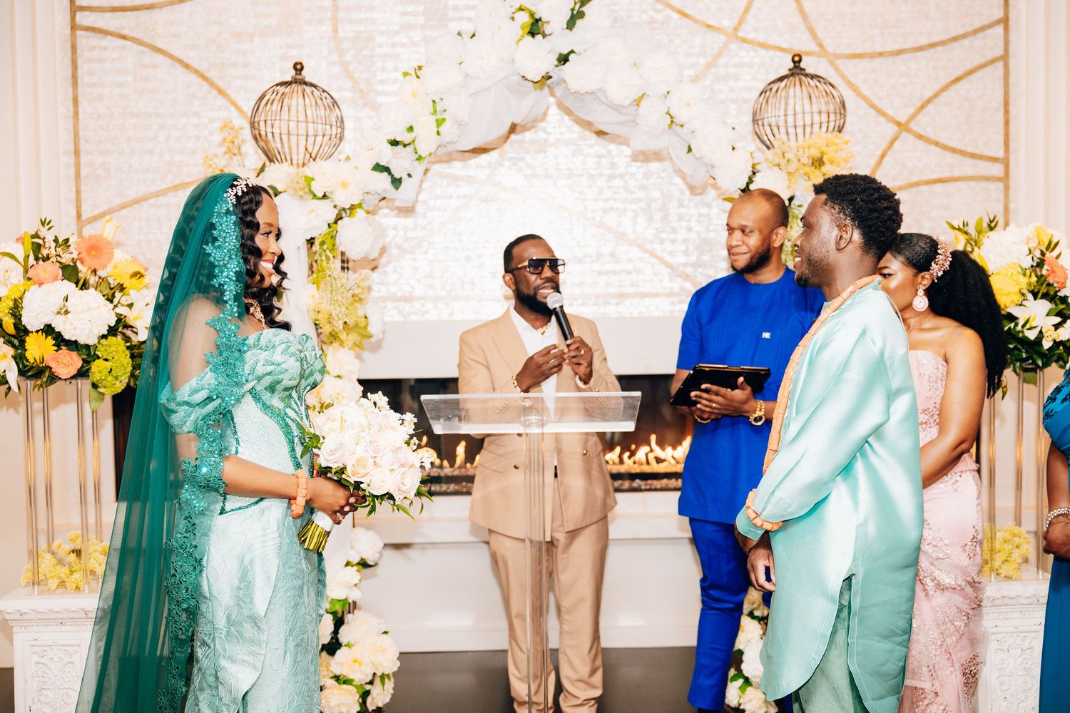 Bride in teal gown and groom in mint suit exchange vows at elegant wedding ceremony with floral arch.