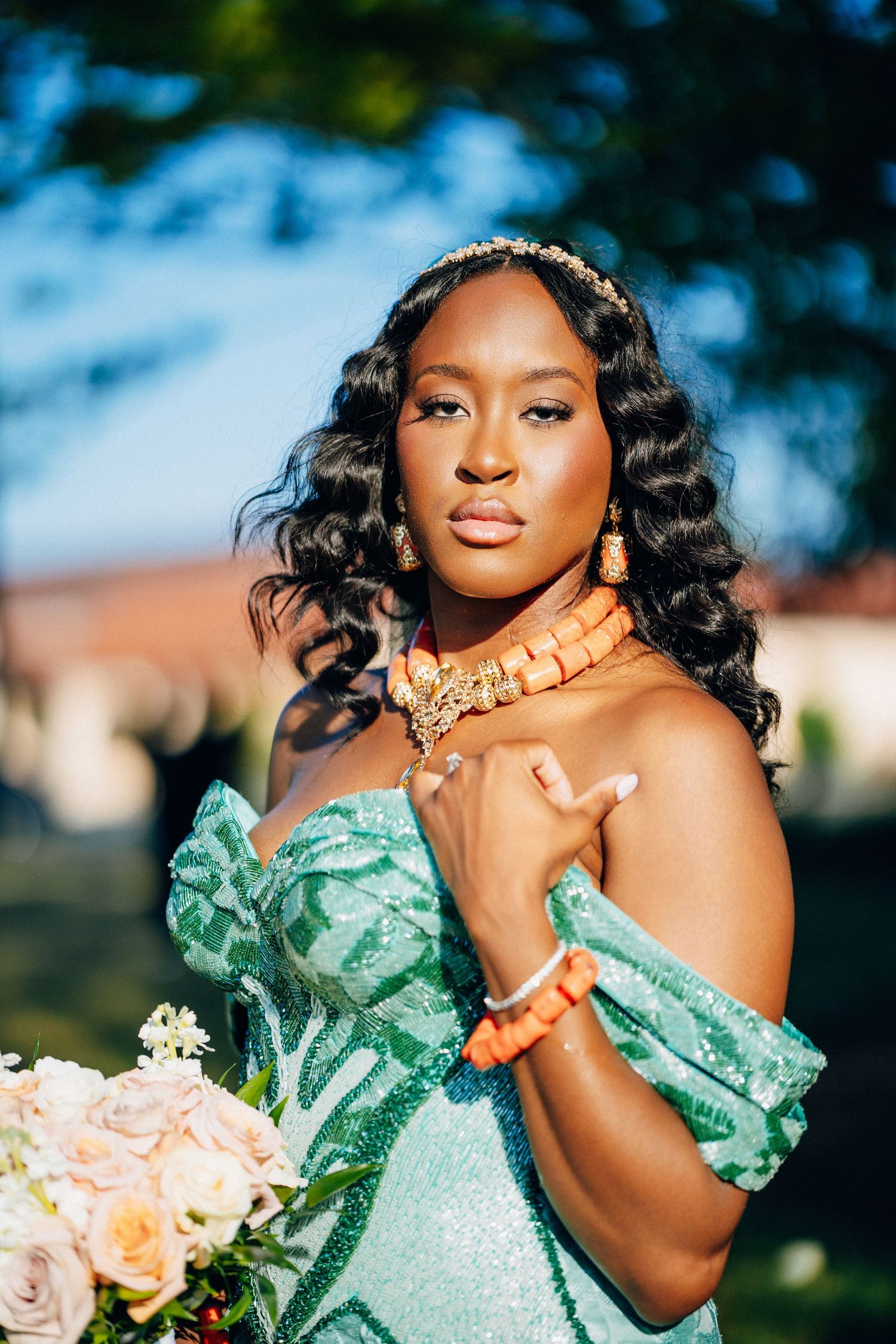 Nigerian bride in mint green gown wearing coral orange jewelry holds white bouquet in outdoor portrait.