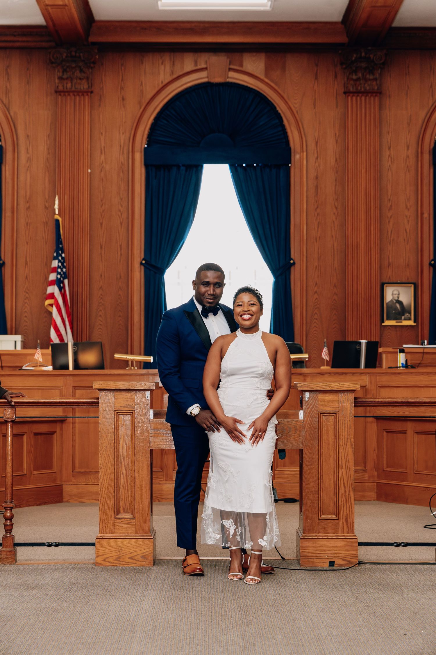 A newlywed couple poses together in a formal courthouse chamber with an American flag and arched woodwork.