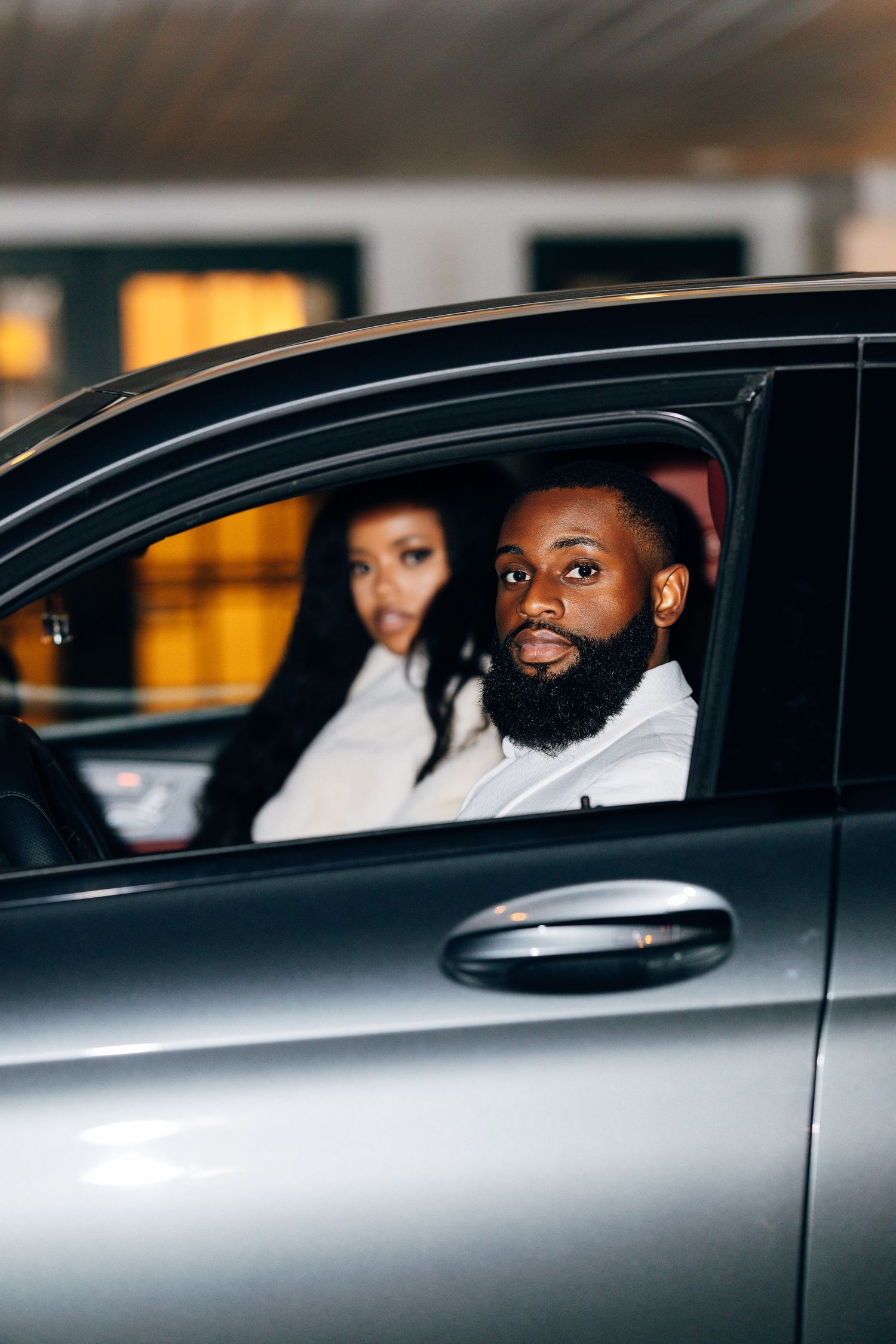 A bearded Black man in a white shirt sits in a silver car with a woman in the passenger seat behind him.
