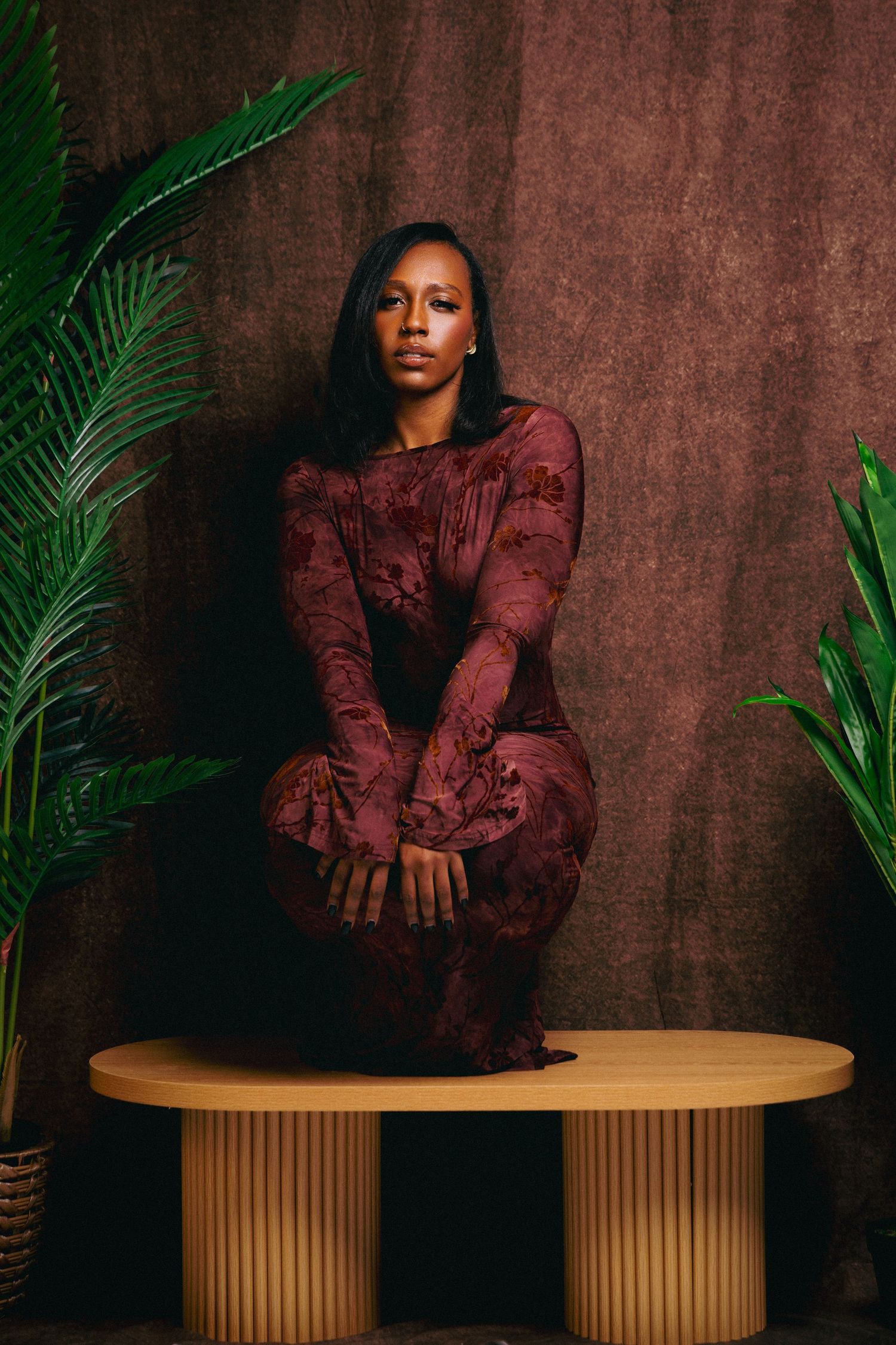 Woman in burgundy floral dress seated on modern wooden table with tropical plants against brown backdrop.