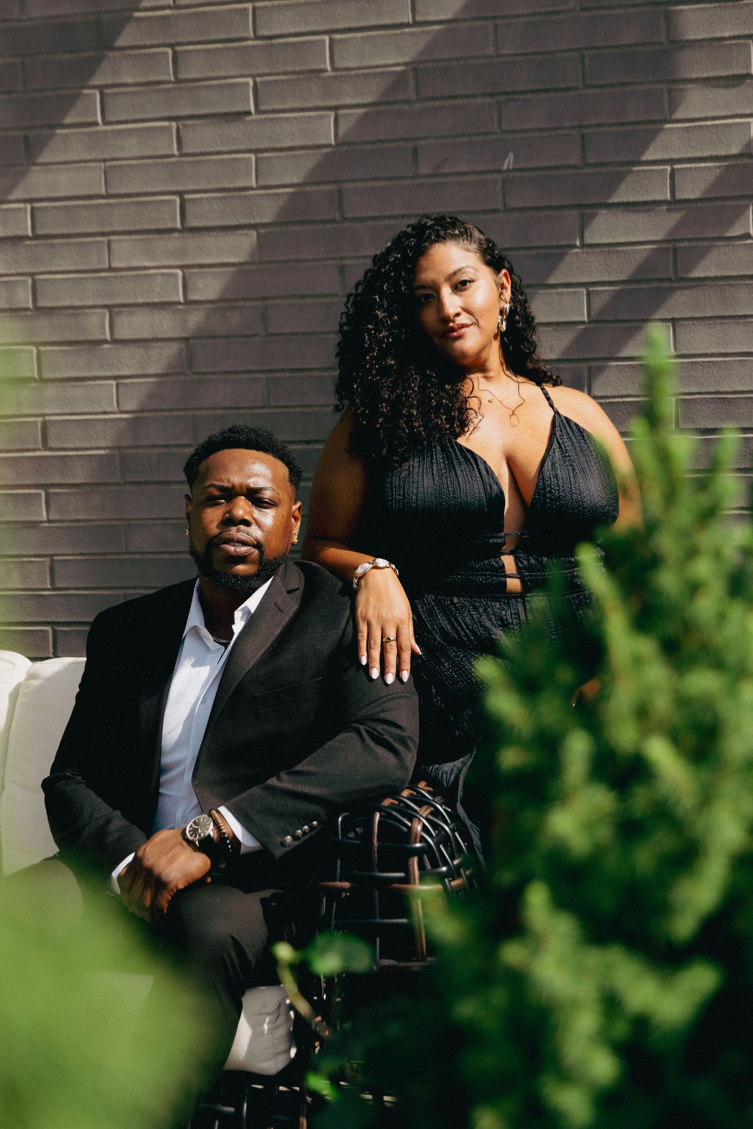 A well-dressed couple poses outdoors against a brick wall, man in black suit seated, woman in black dress standing beside him.