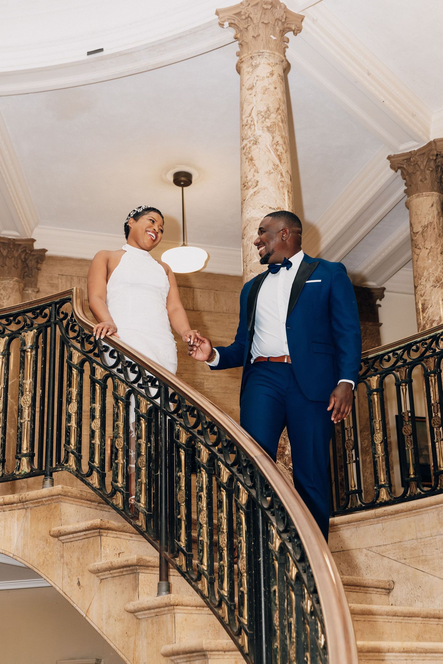 Bride and groom hold hands smiling on curved marble staircase with ornate iron railing.