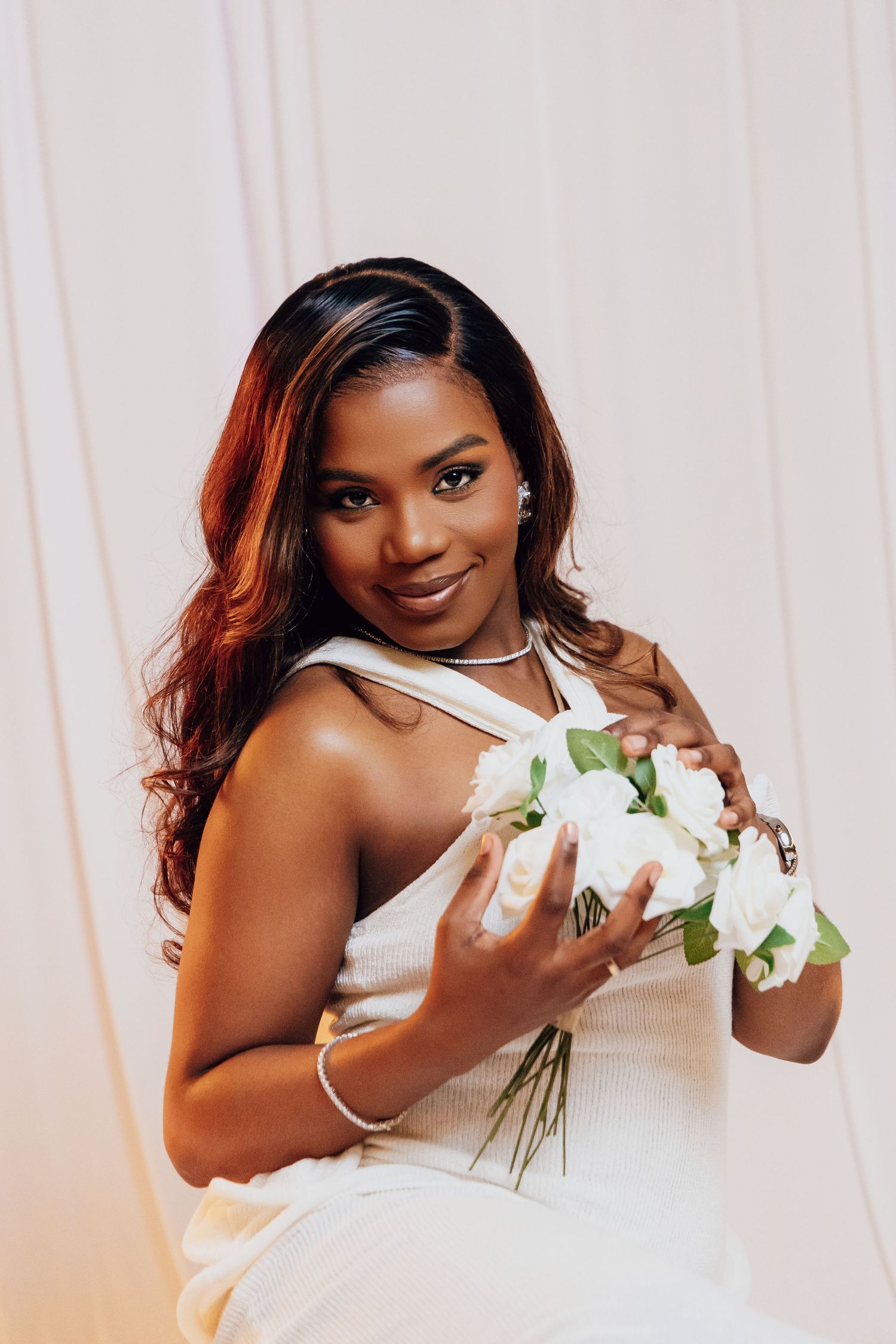 Elegant Black woman in white dress holding bouquet of white roses, smiling gracefully for a bridal portrait.