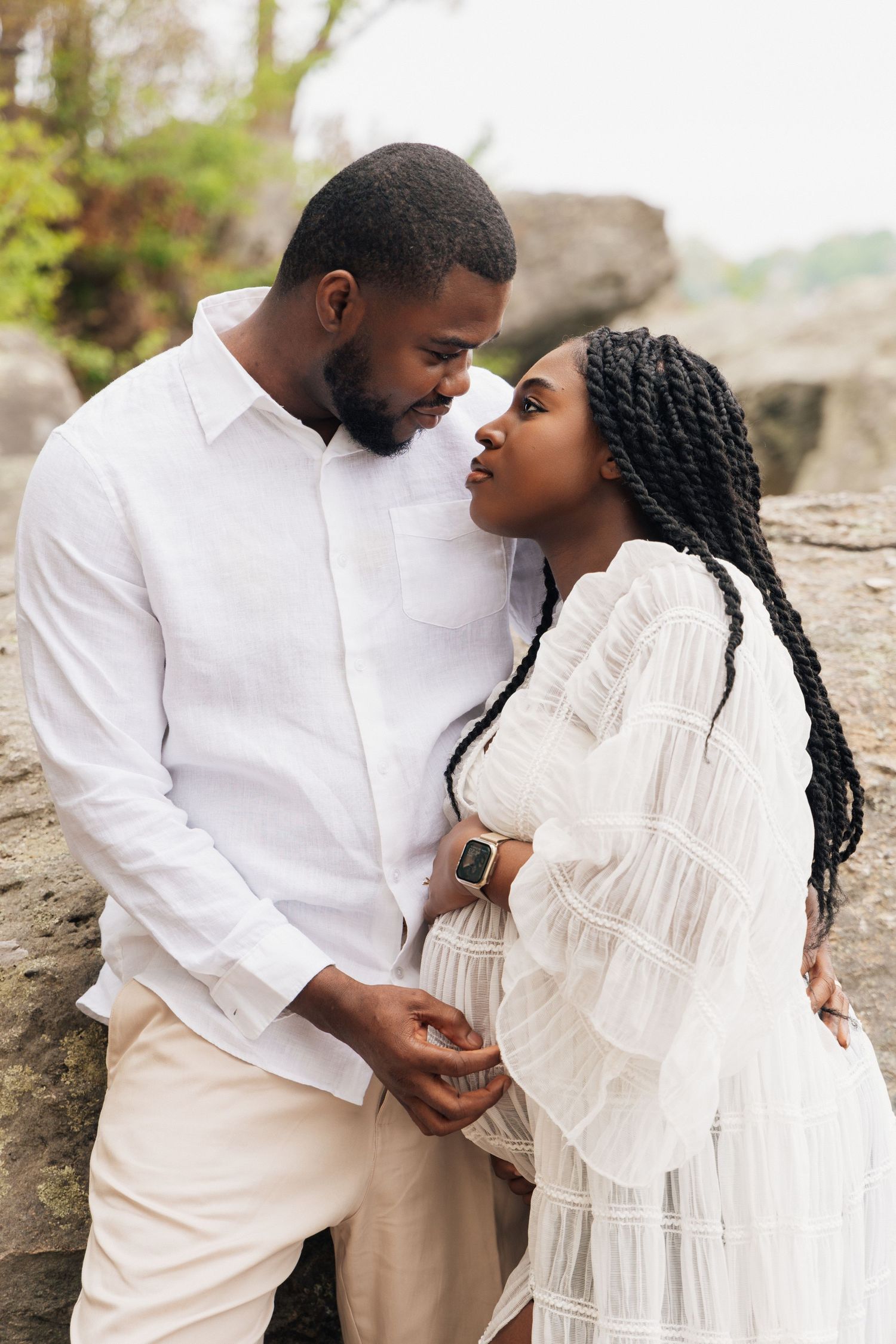 A couple in white outfits share a tender moment outdoors, with the woman visibly pregnant, holding hands near rocky scenery.