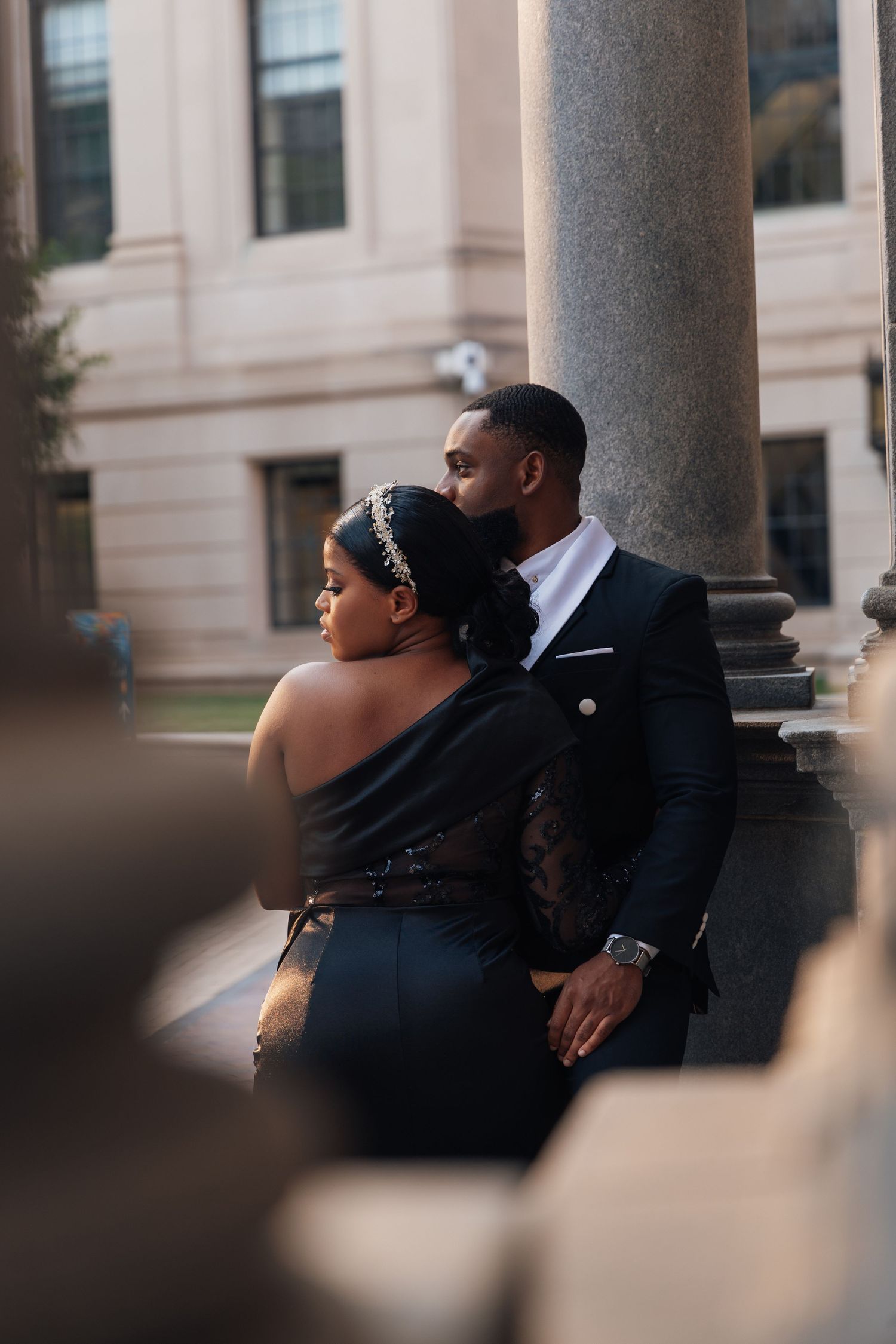 Elegant Black couple in formal attire pose near stone columns at a classic building during golden hour.