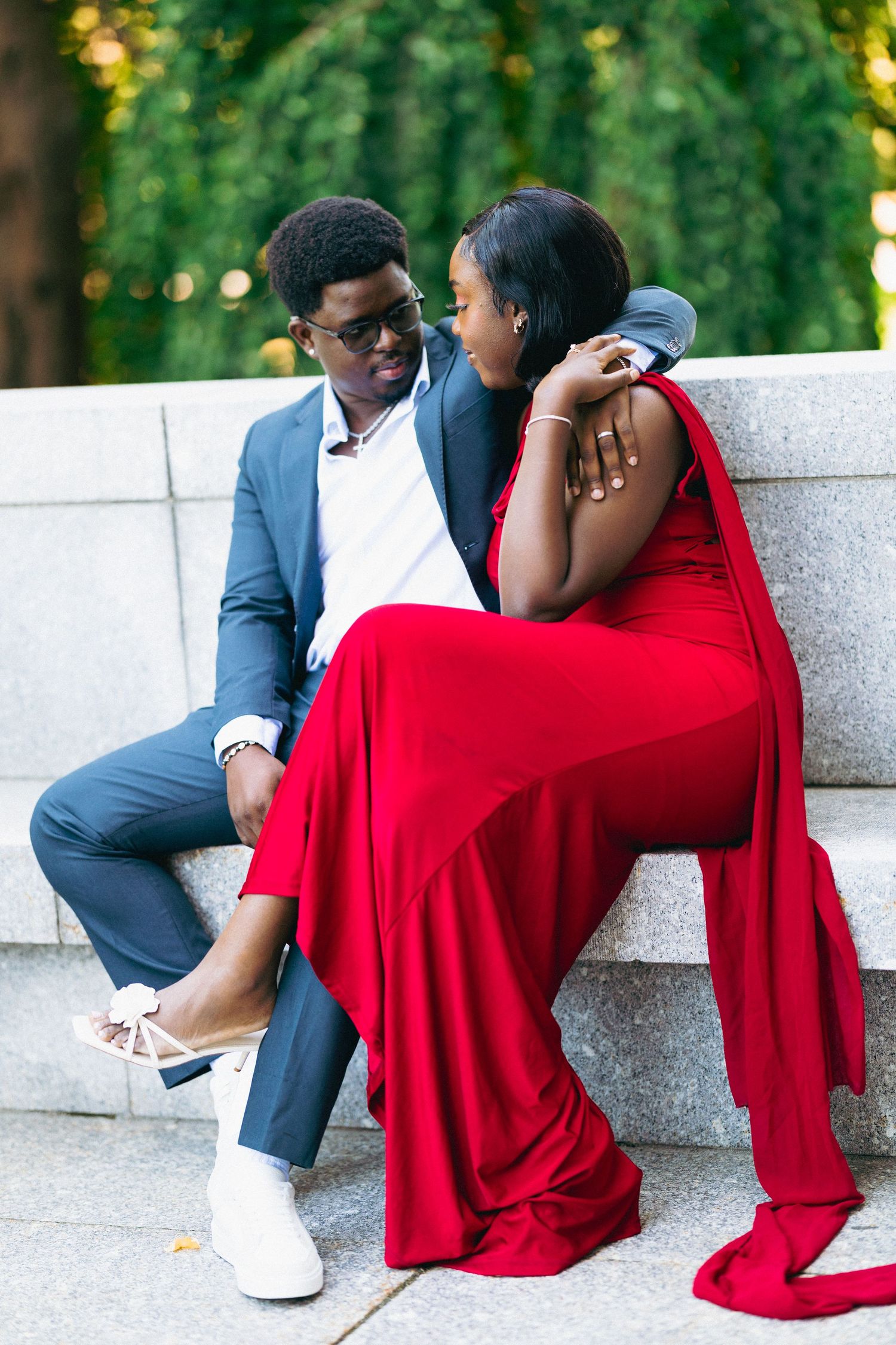 A couple in elegant attire, man in gray suit and woman in red gown, share a romantic moment on stone steps.