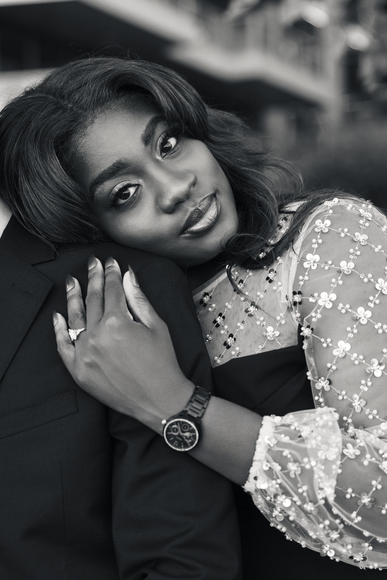 Black and white portrait of a smiling woman in floral lace top wearing a watch, embracing a man in a suit.