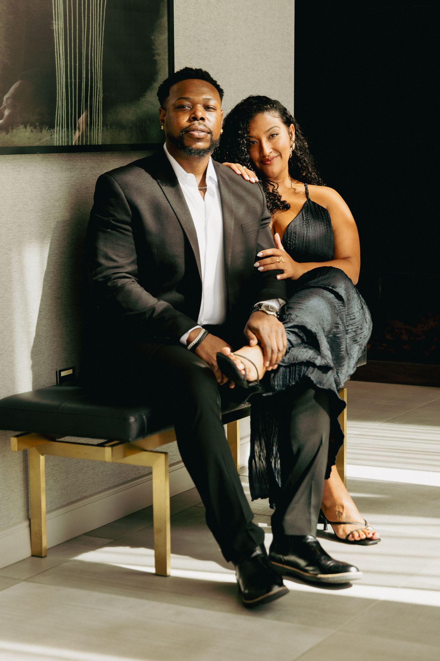 Elegant couple posing together, man in black suit and woman in black dress, seated in a sunlit modern interior.