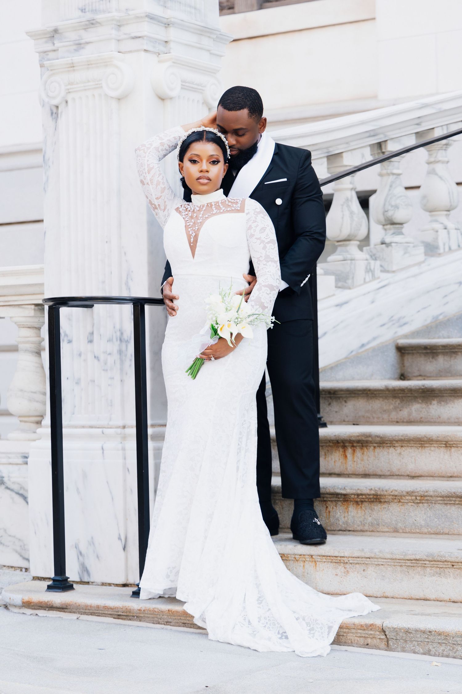 Elegant Black couple in wedding attire pose on marble steps, bride in lace gown holding white bouquet, groom in black tuxedo.