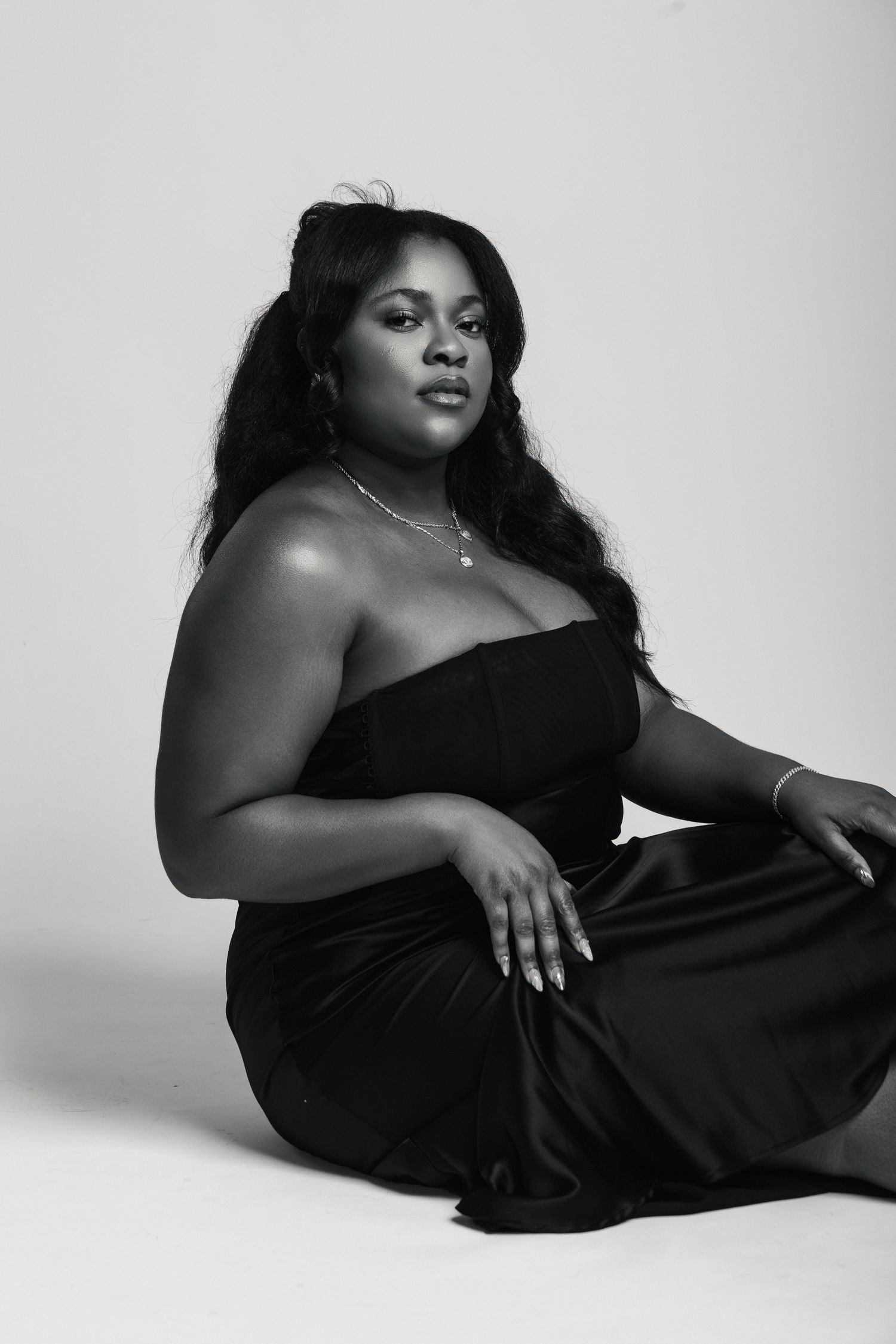Black and white photo of a woman in a strapless black dress sitting gracefully on the floor.
