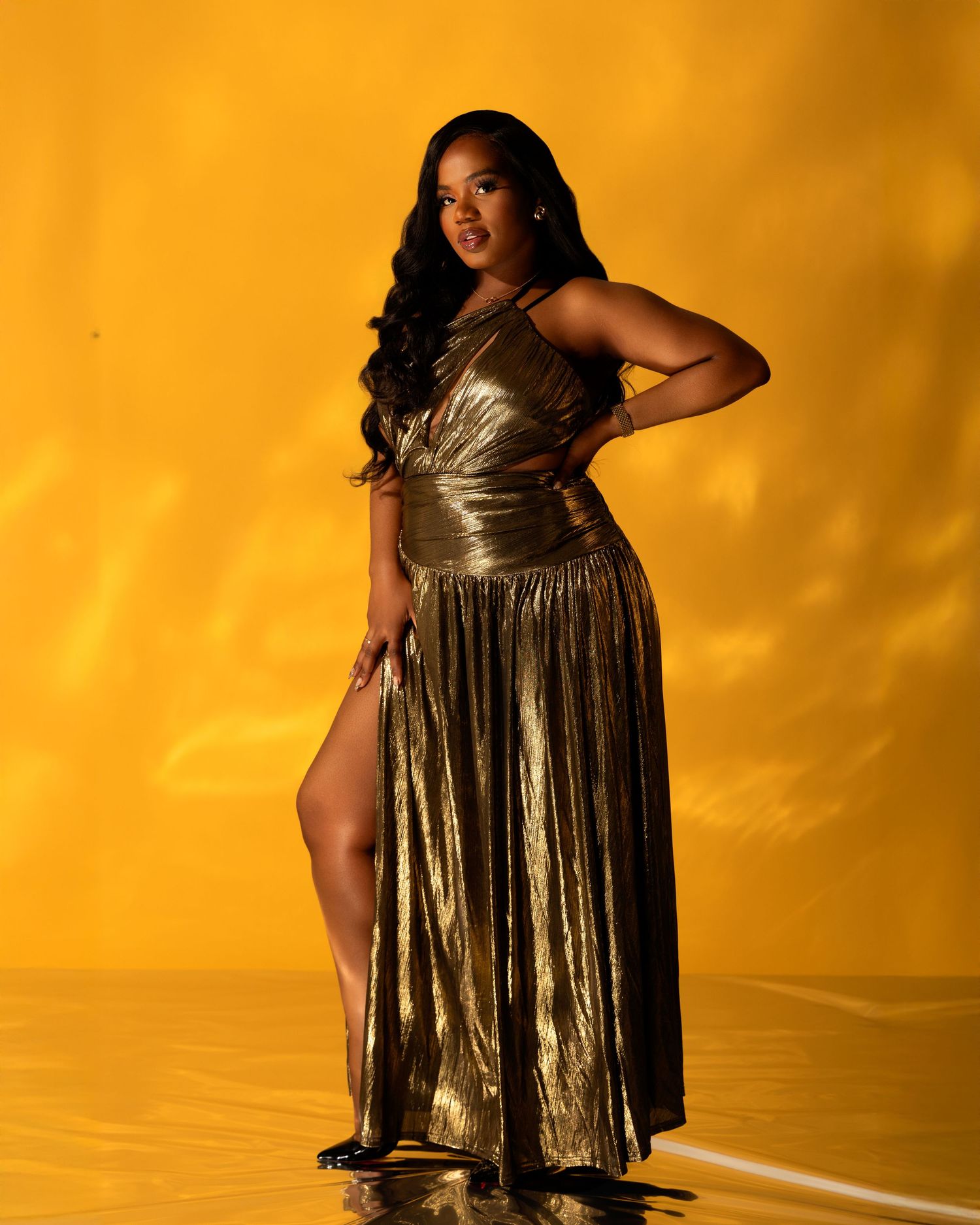 Woman in gold metallic gown poses confidently against vibrant yellow background in elegant fashion portrait.