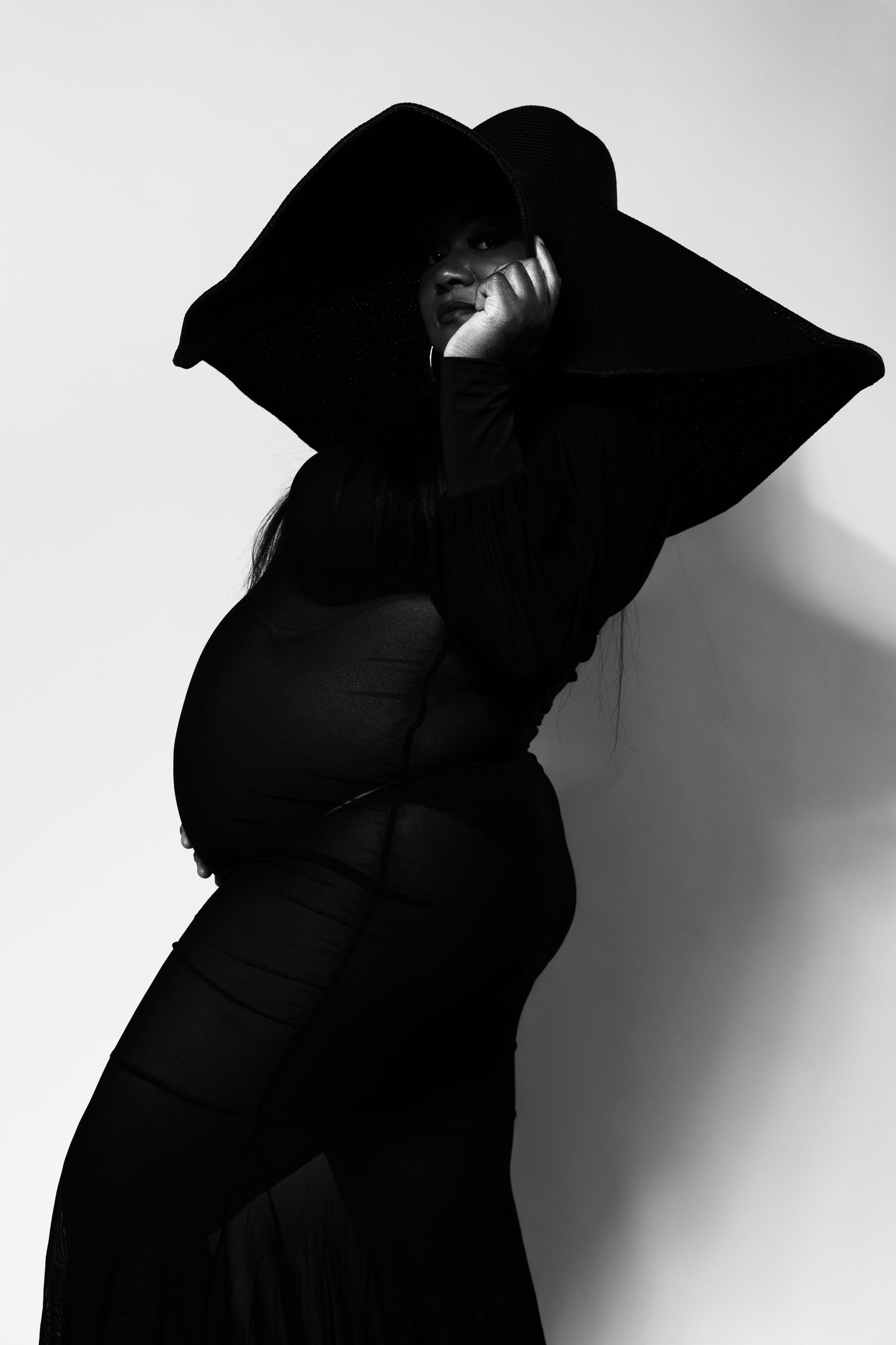 Black and white photo of a pregnant woman in a wide-brim hat and dark dress, posing dramatically against a light background.