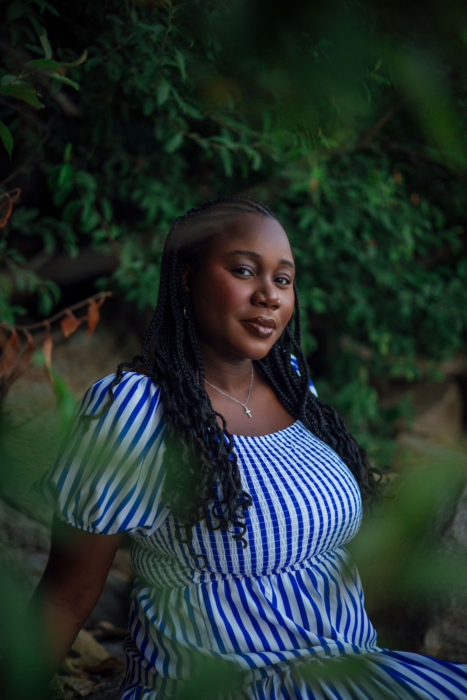 Young Black woman with braids in blue striped dress poses outdoors among lush green foliage.