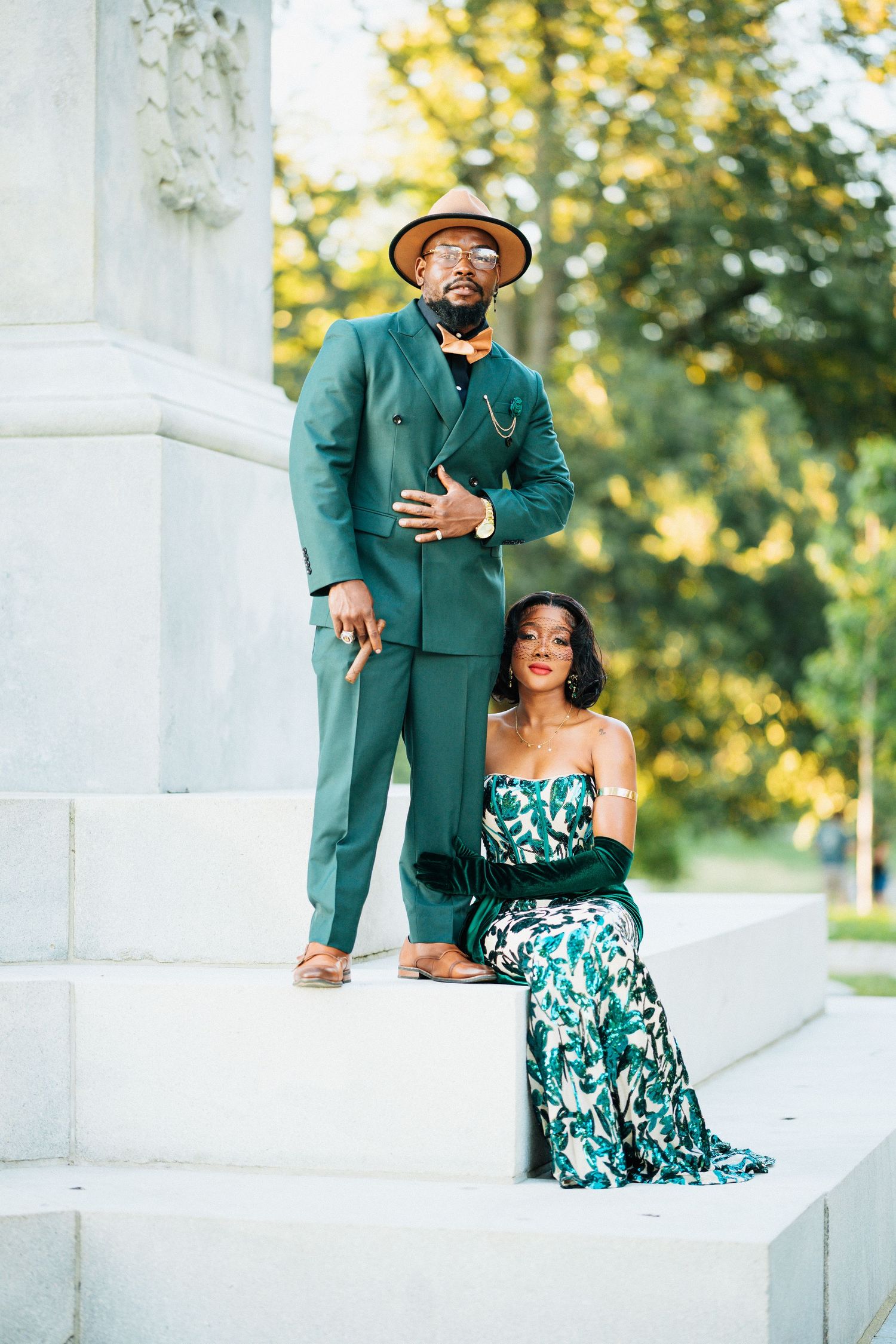 Elegant couple posing on white marble steps, man in green suit with hat, woman in floral green gown.