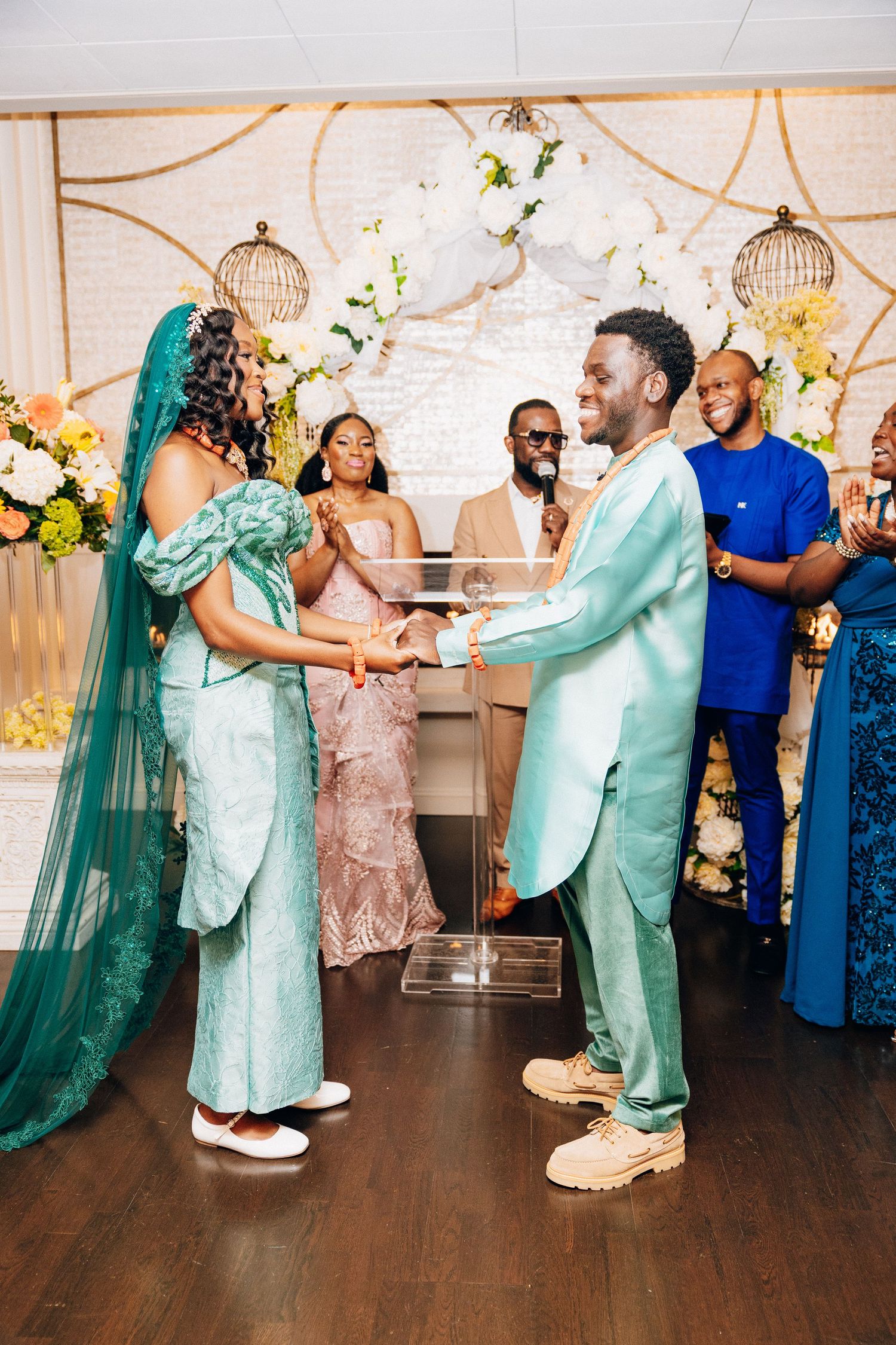 Couple in matching mint green outfits exchange vows at elegant wedding ceremony with floral backdrop and guests applauding.