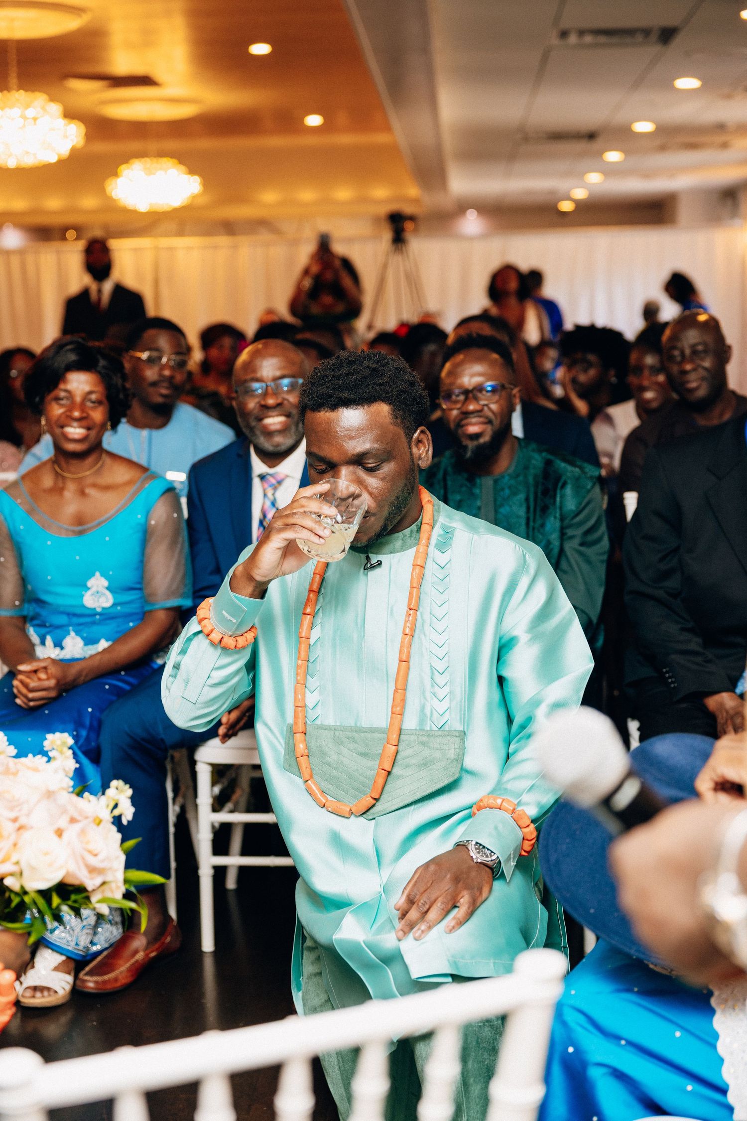 Groom in teal African attire drinks from cup during traditional wedding ceremony.
