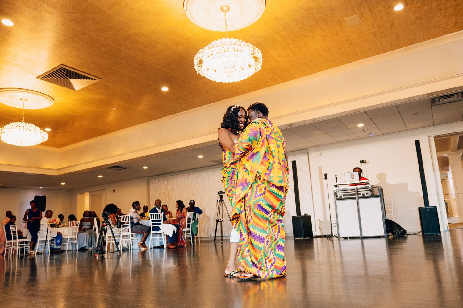 Couple sharing a first dance in a golden-ceilinged ballroom wearing colorful traditional Kente cloth attire.