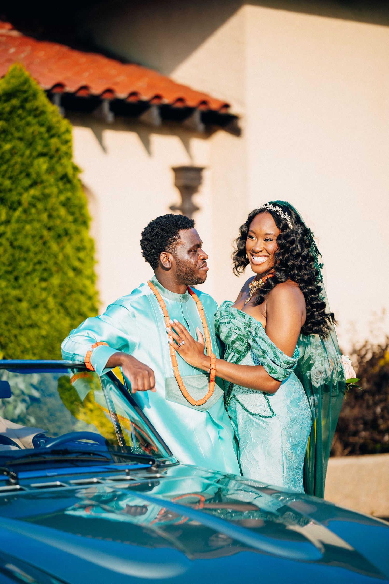 Nigerian wedding couple in matching teal traditional attire laughing together beside a vintage blue convertible car.