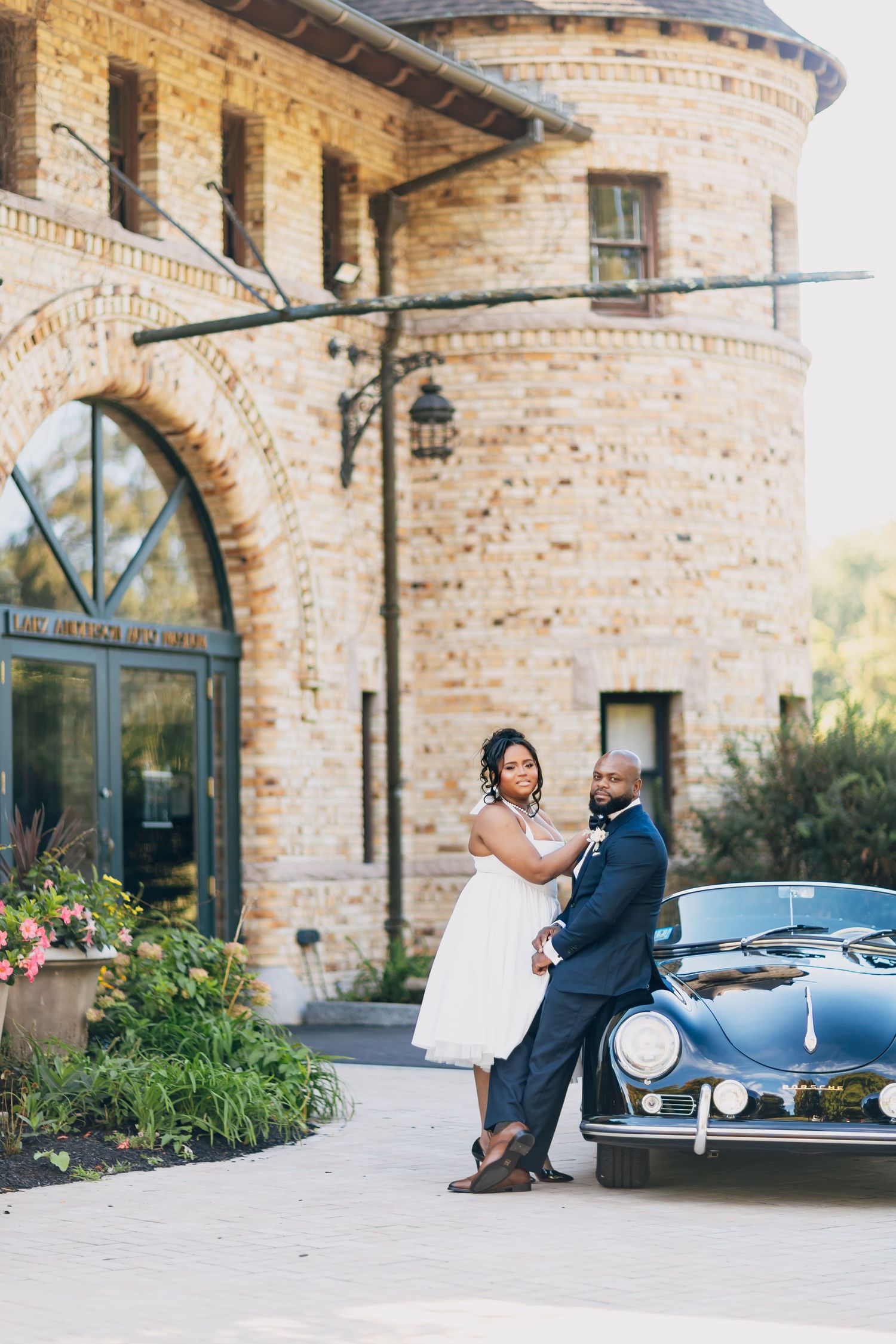 Elegant couple in wedding attire lean against a classic black car before a historic brick building.