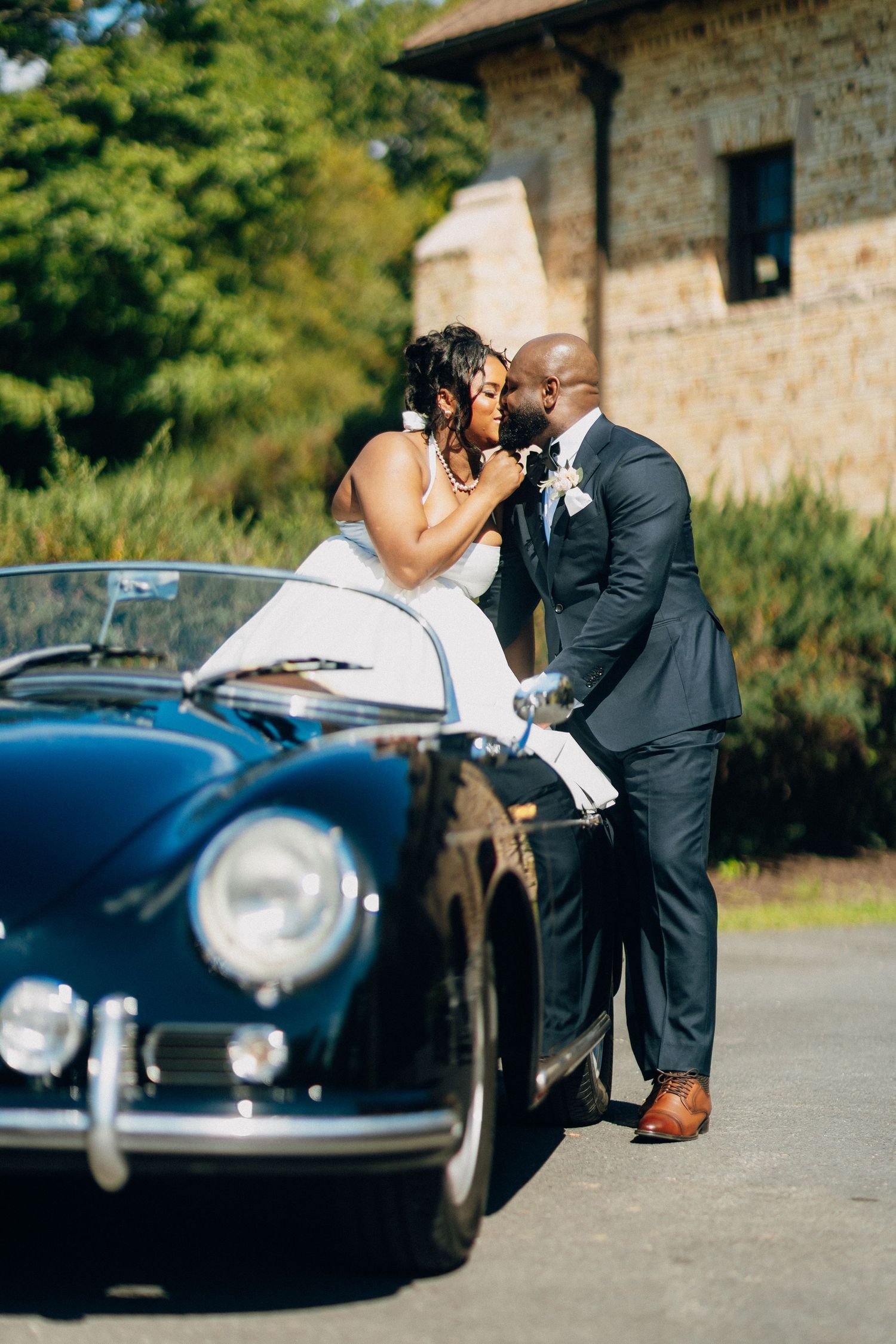 Black bride and groom sharing a kiss beside a vintage navy Porsche convertible on their wedding day.