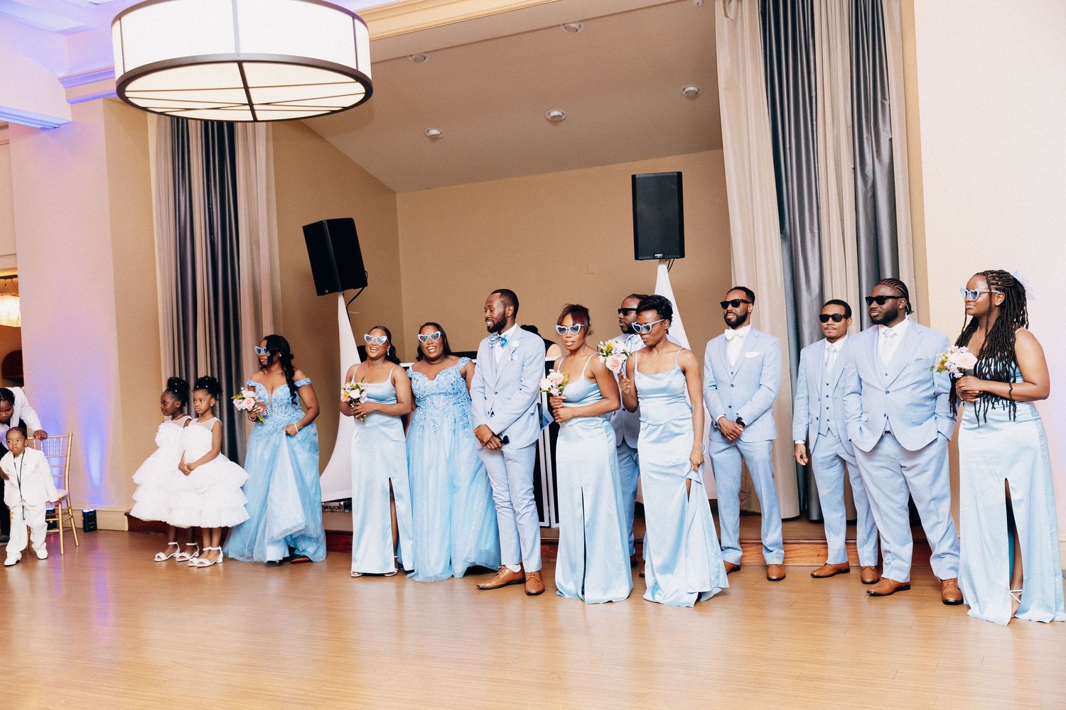Wedding party in light blue attire poses together on a dance floor during a elegant indoor wedding reception.
