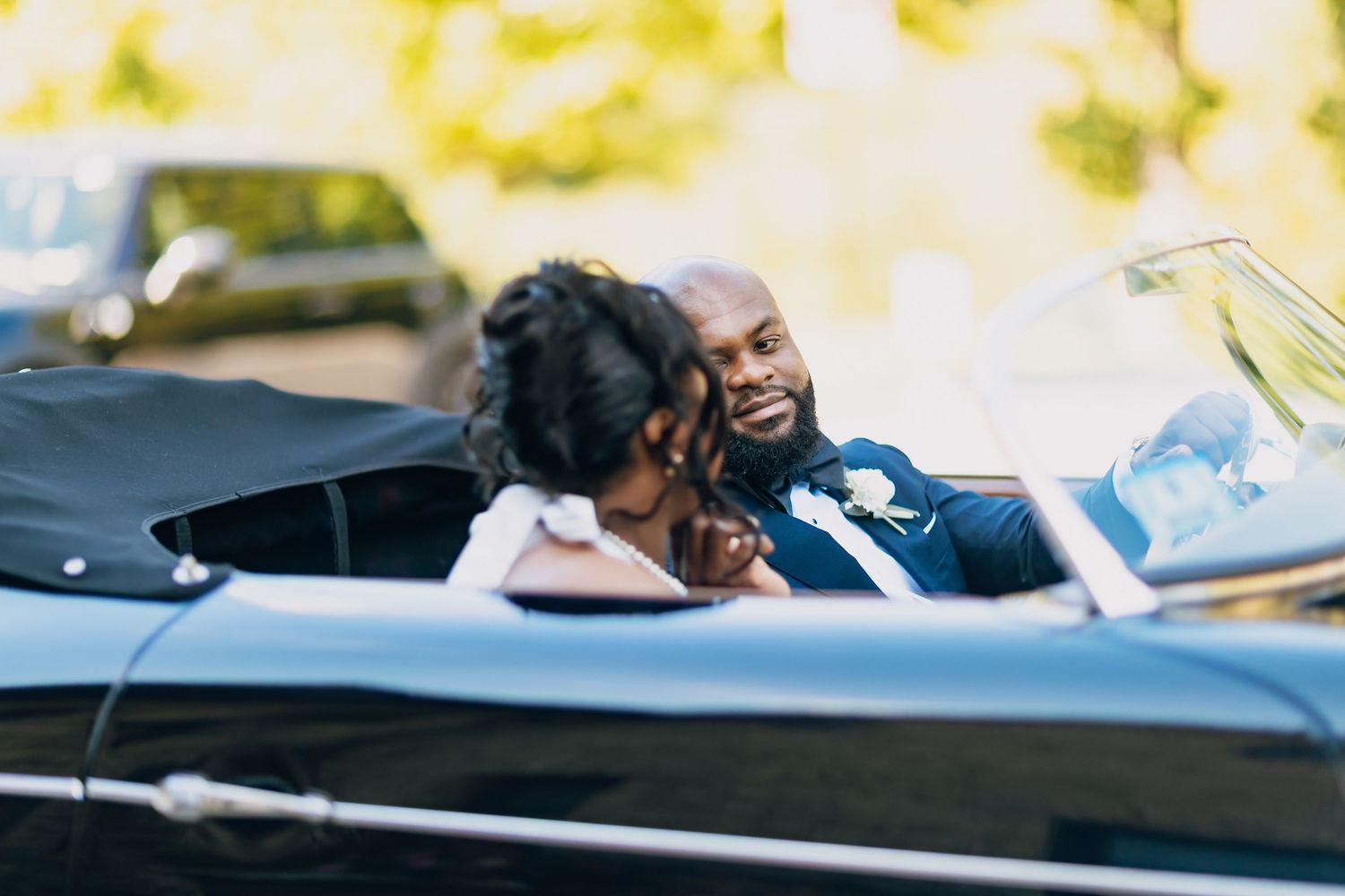 Black bride and groom smiling at each other while seated in a classic black vintage convertible car.