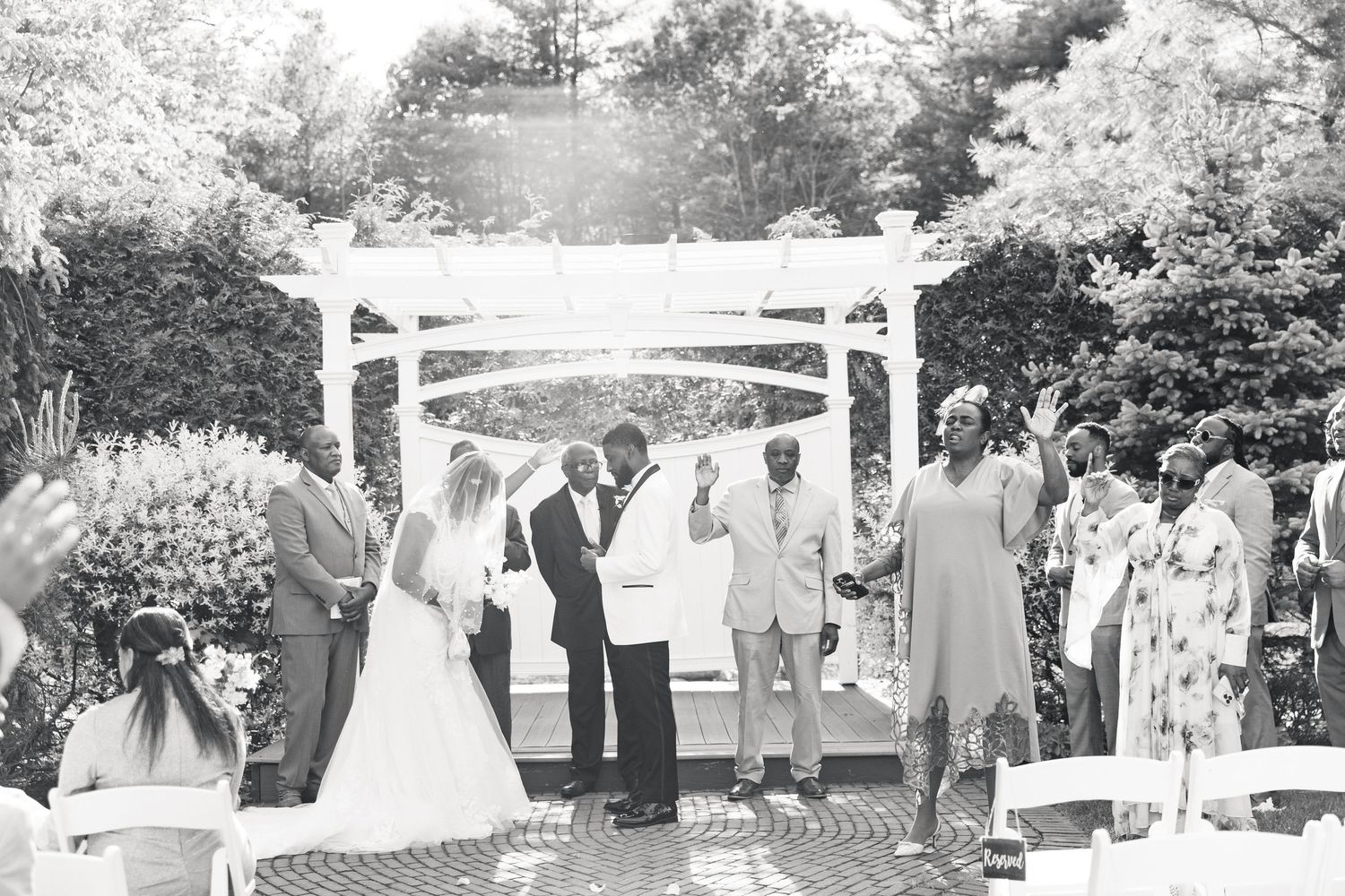 Bride and groom celebrate at outdoor wedding ceremony under white pergola with guests cheering around them.
