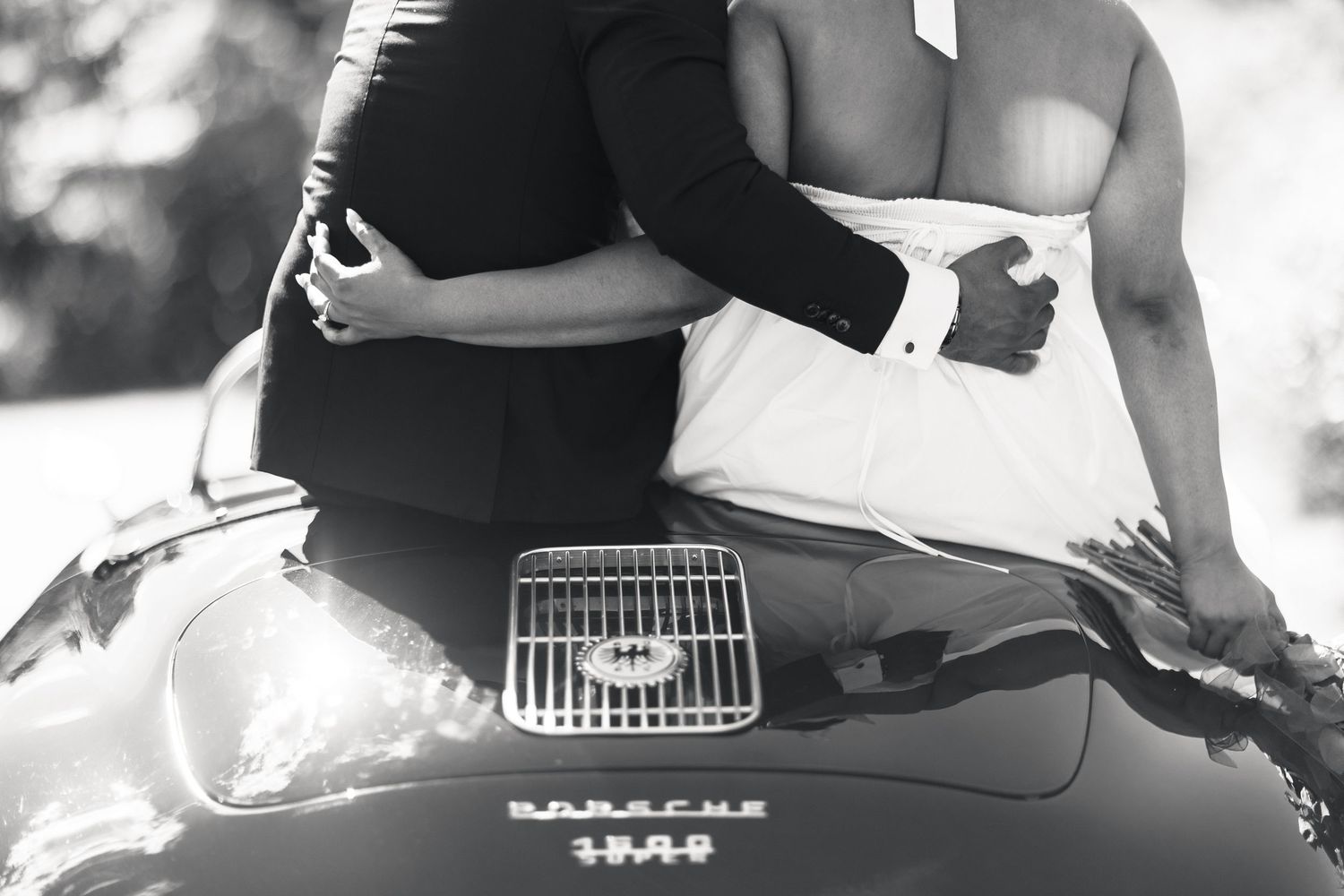 Black and white close-up of bride and groom embracing on hood of vintage Porsche 356 convertible.