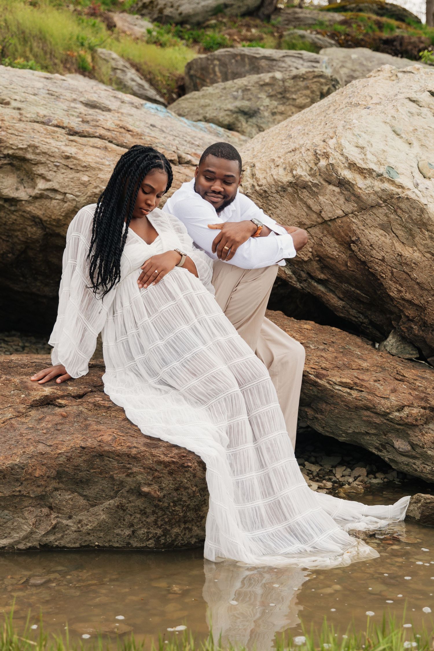 A pregnant couple in white outfits sit on rocks near water, the man tenderly embracing the woman from behind.