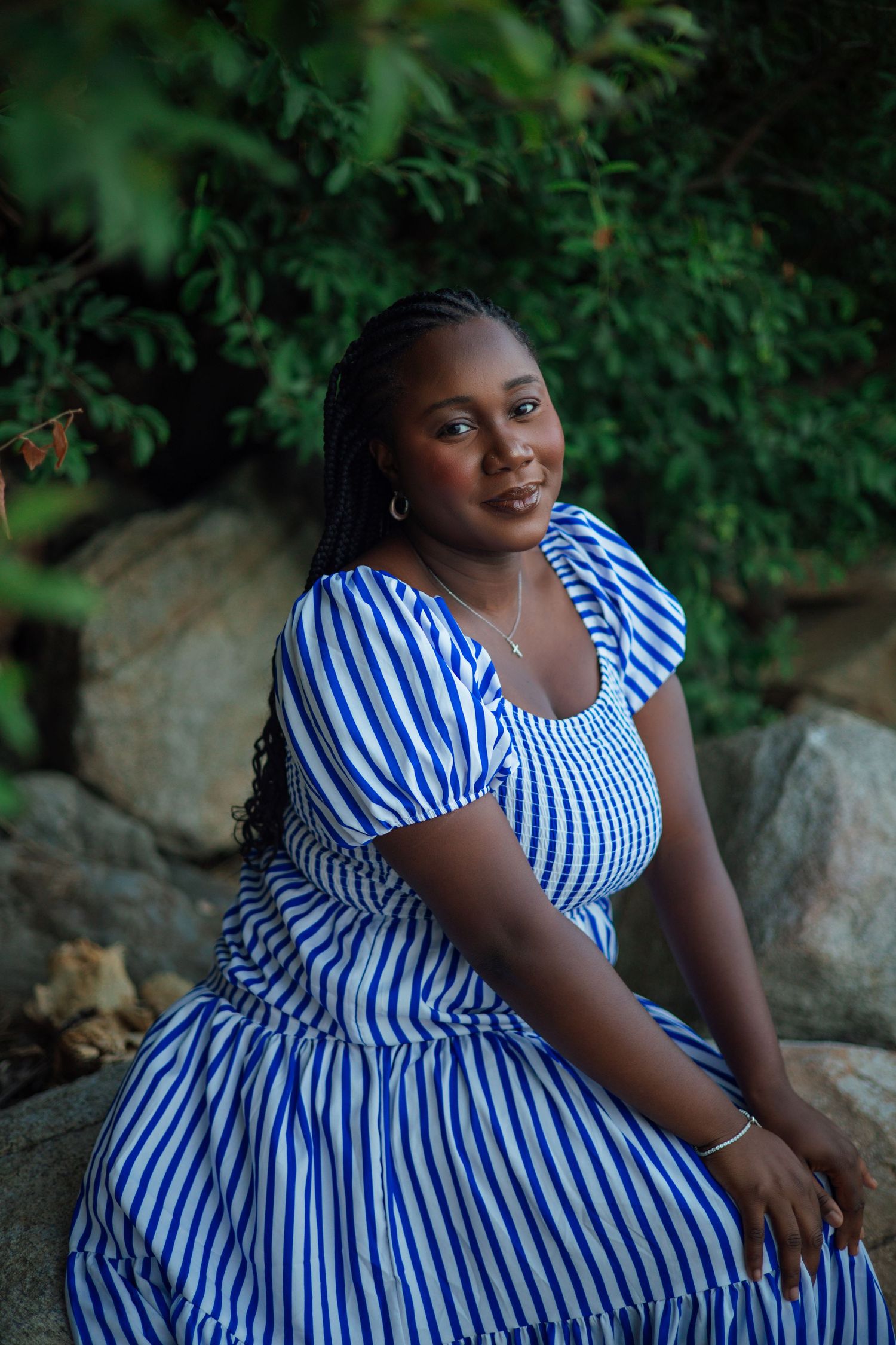 Young woman in blue striped dress sitting on rocks surrounded by lush green foliage, smiling warmly.