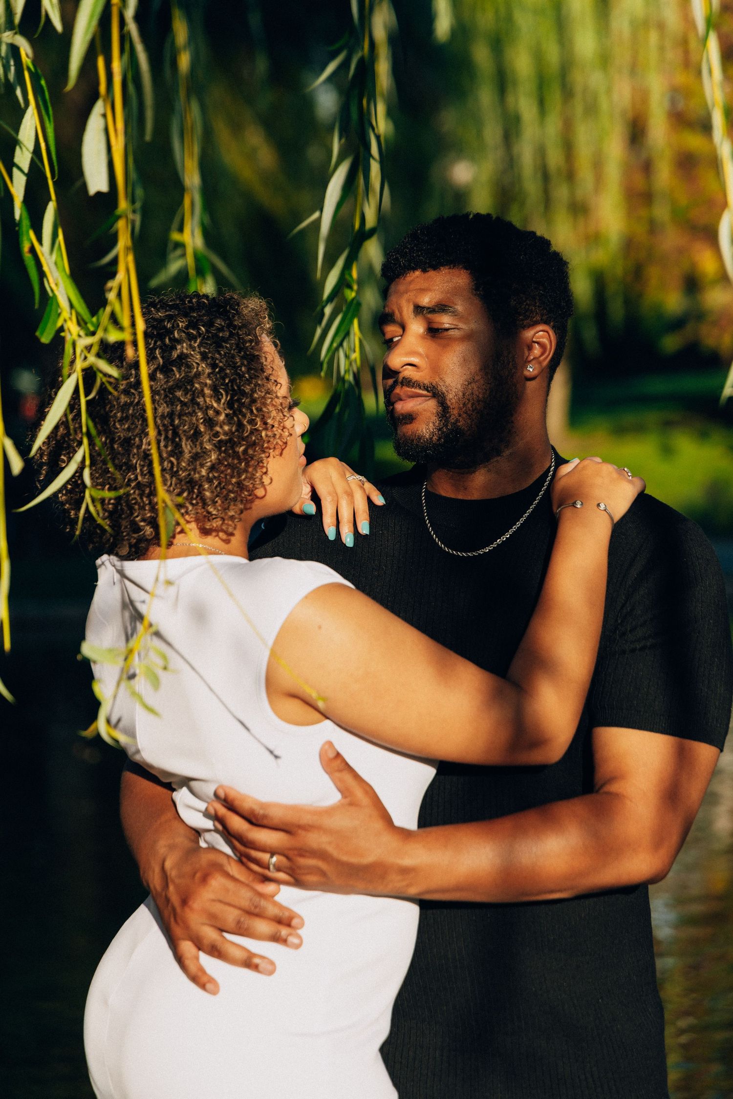 An affectionate Black couple slow dancing under weeping willow branches in warm golden sunlight in a lush park setting.