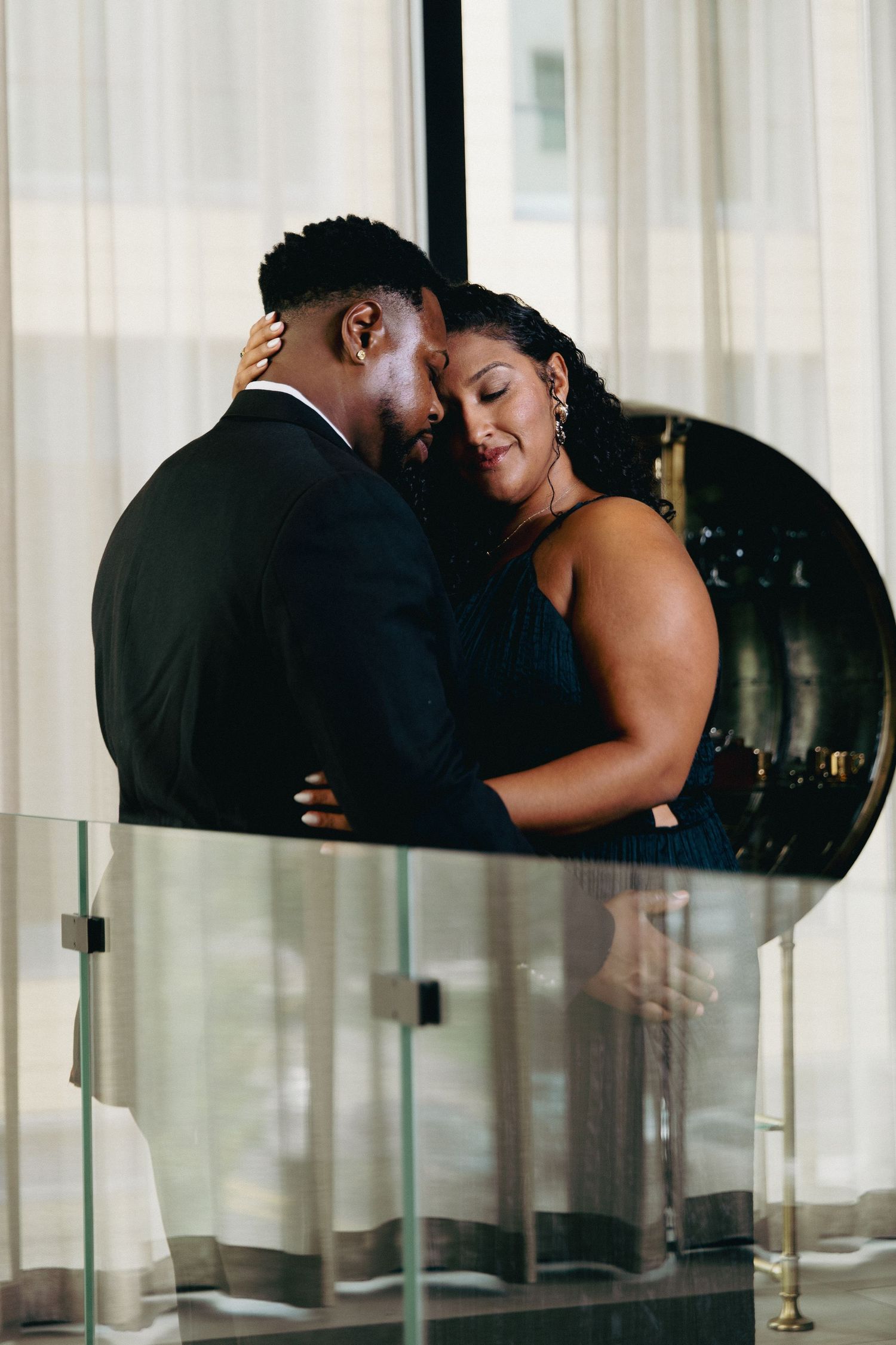 Elegant couple in formal attire sharing an intimate embrace near a glass railing with sheer curtains backdrop.