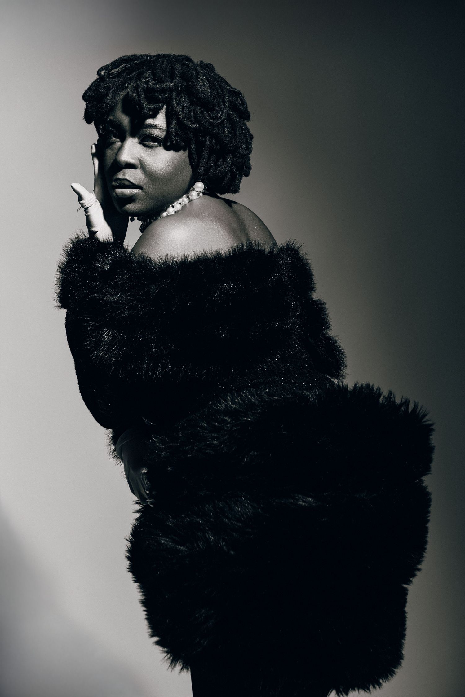 Black and white portrait of a woman in a black fur coat and pearl necklace posing elegantly against a gray background.