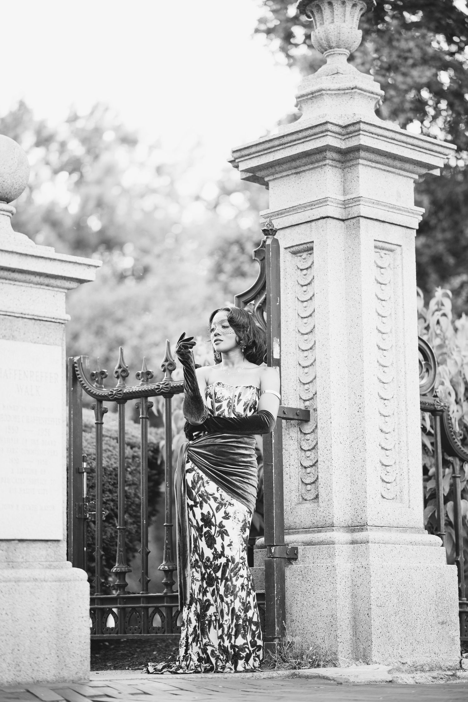 Black and white photo of elegant woman in floral gown with green gloves posing at ornate iron gates.