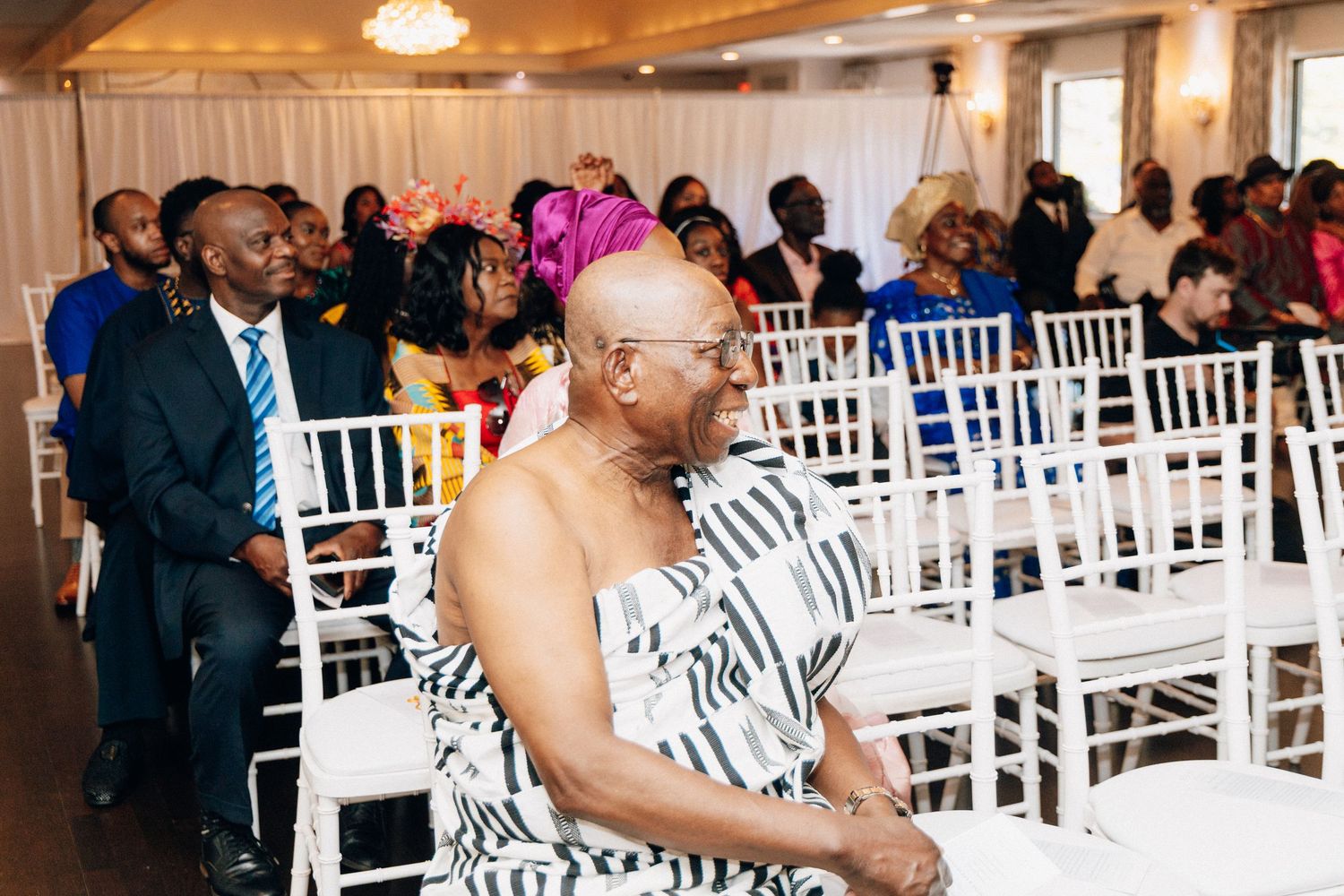 Guests seated at an elegant indoor wedding ceremony, with a man in traditional Kente cloth attire in the foreground.