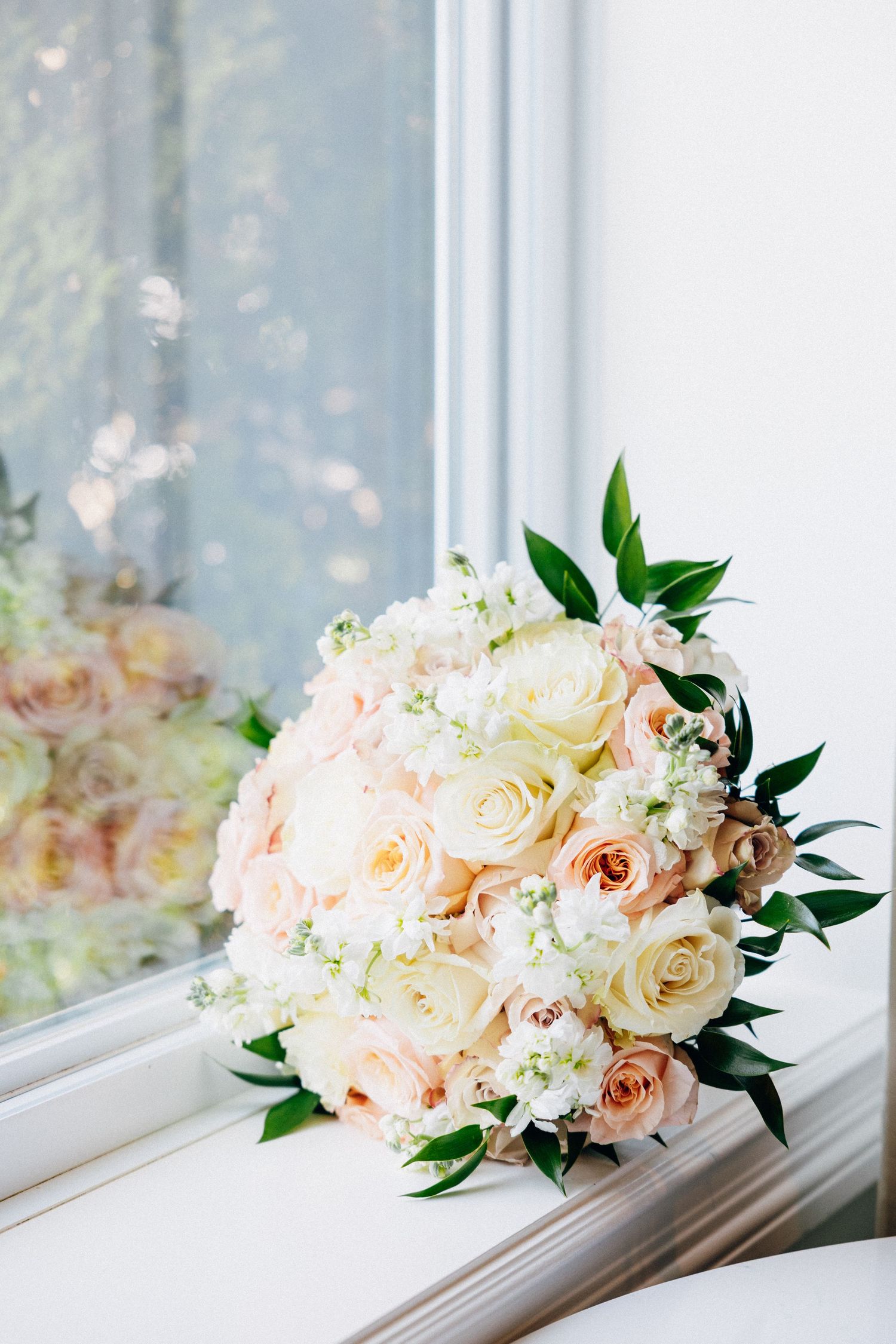 Elegant bridal bouquet of blush and cream roses with greenery resting on a white windowsill.