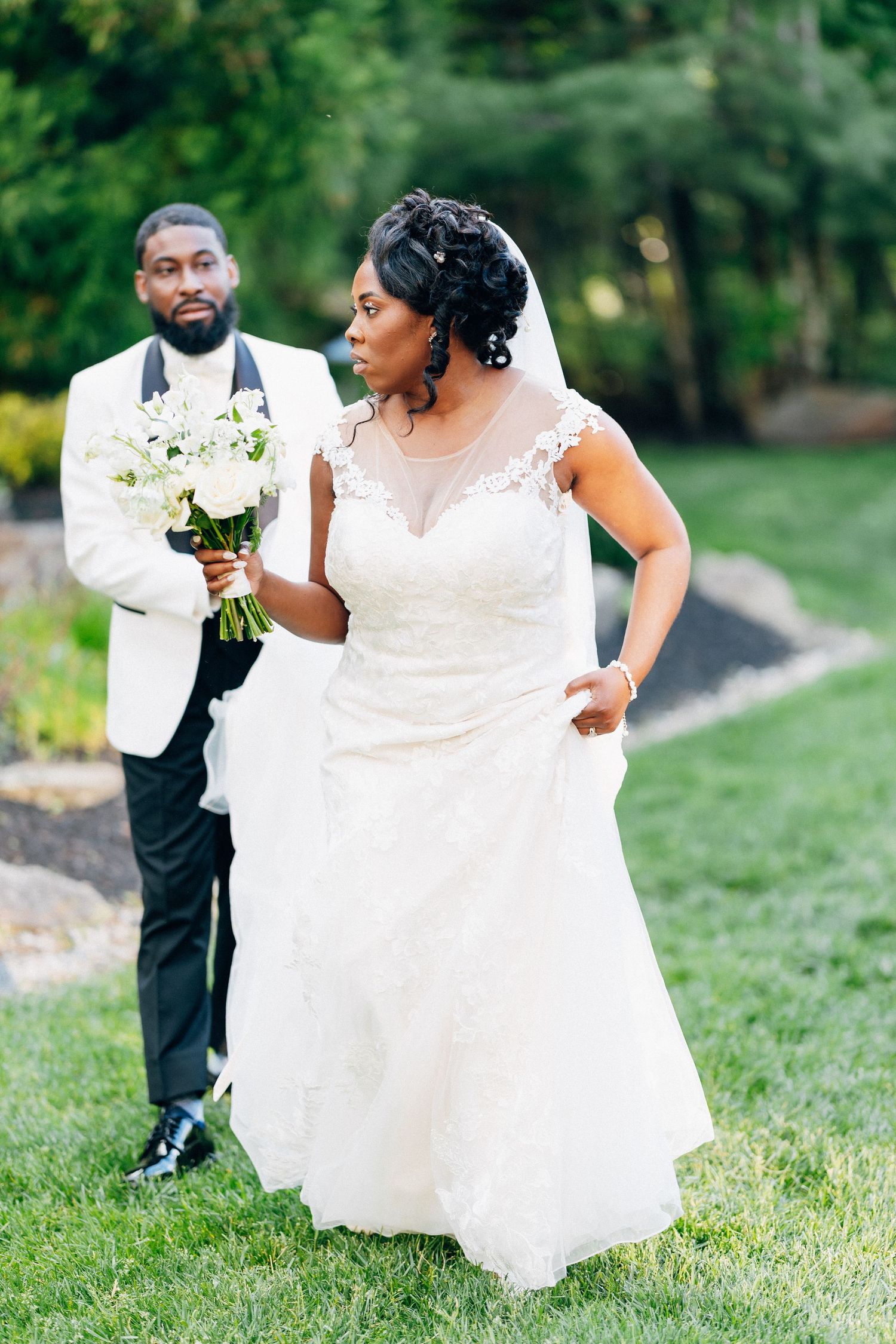 A bride in lace gown holds white bouquet while groom in white tuxedo jacket stands behind her on green lawn.