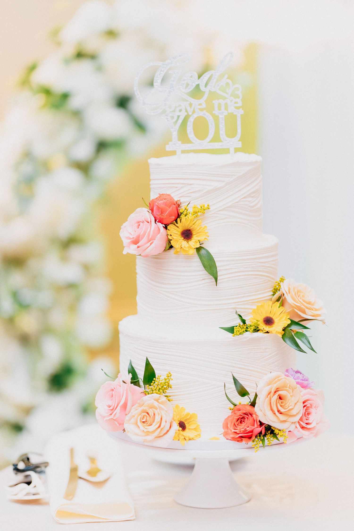 Elegant three-tier white wedding cake adorned with colorful roses and sunflowers, topped with a decorative cake topper.