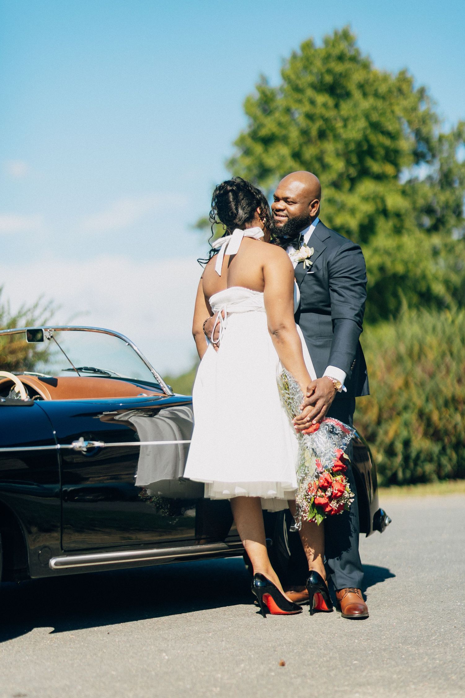 Newlyweds sharing a romantic moment beside a classic black convertible, bride holding a bouquet.