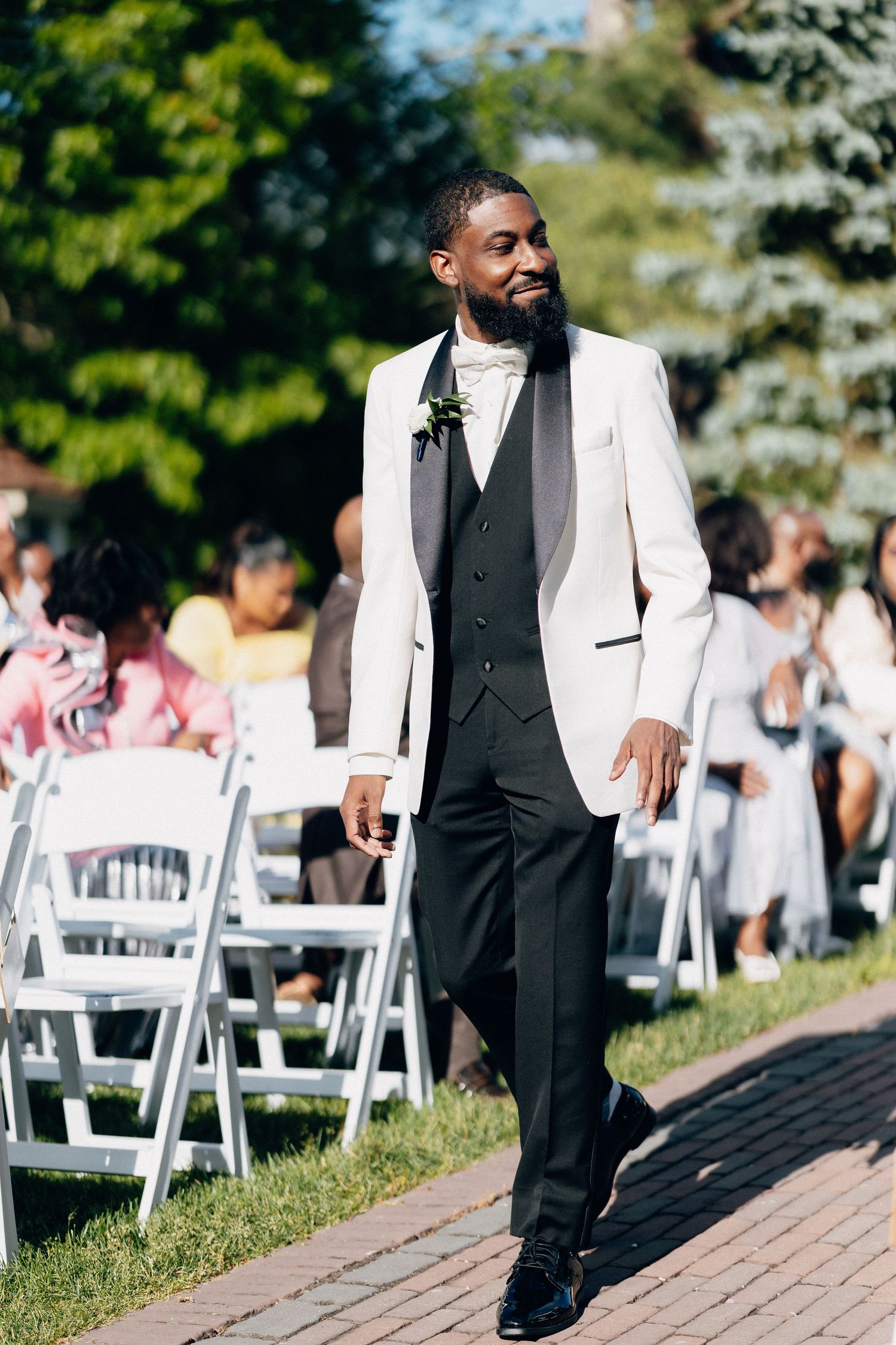 Elegant groom in white tuxedo jacket with boutonniere walking down brick aisle at outdoor wedding.