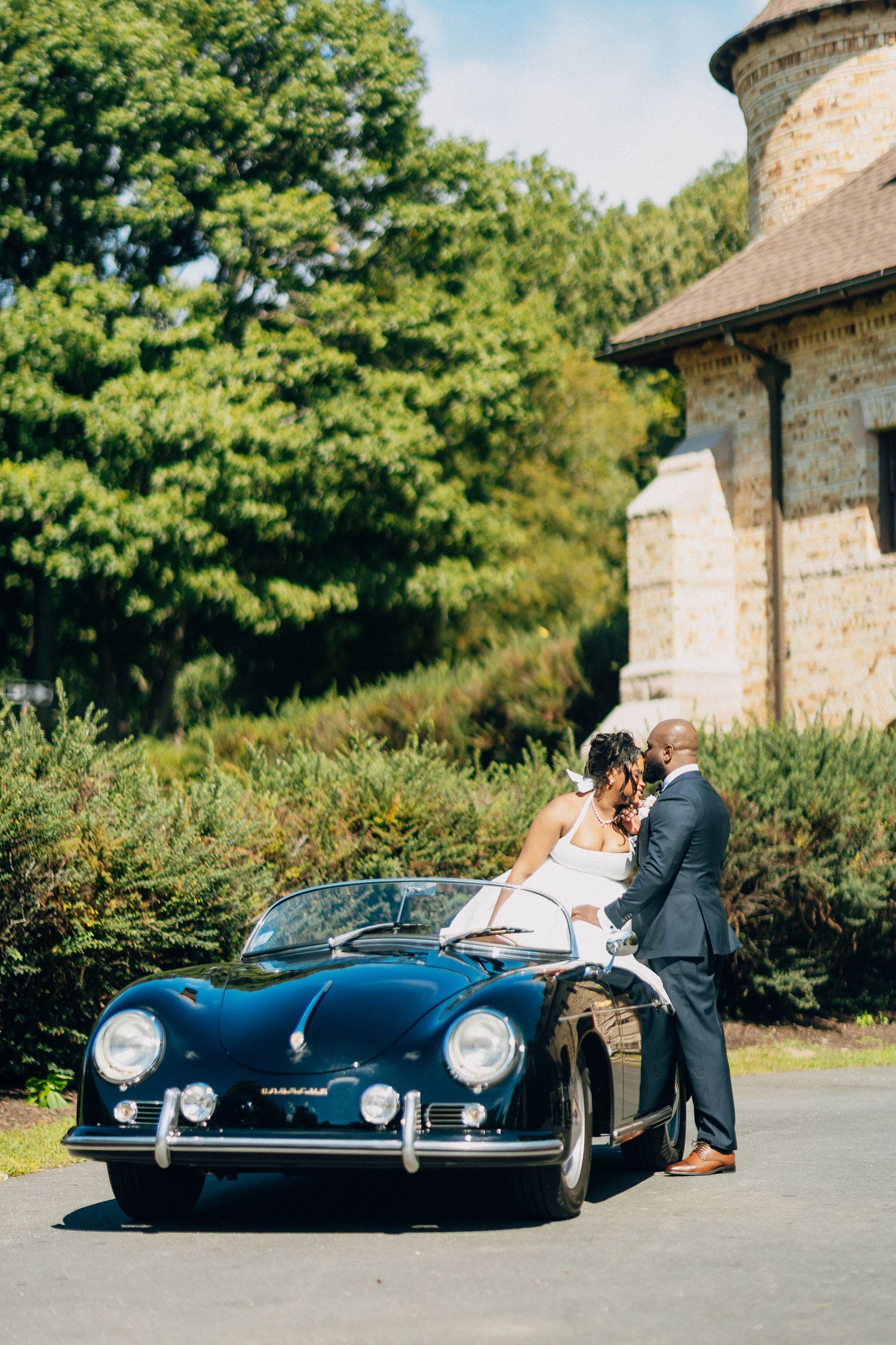 Wedding couple kissing beside a classic navy Porsche 356 convertible outside a stone building.